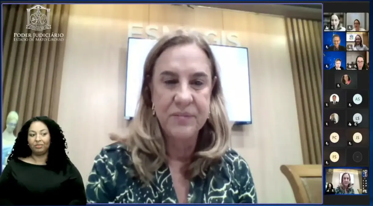 Tela de videoconferência institucional. Ao centro, uma pessoa sentada em ambiente formal, com painel de madeira ao fundo e tela com identidade visual do Poder Judiciário de Mato Grosso. À direita, aparecem miniaturas de outros participantes em colunas verticais