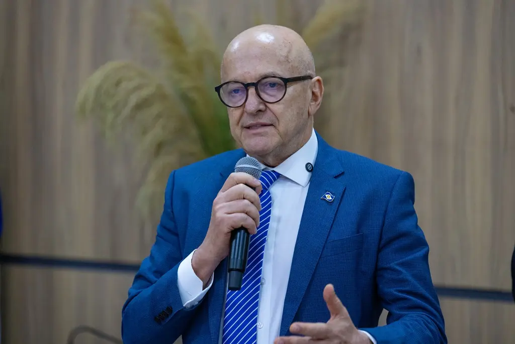 Foto horizontal em plano fechado que mostra o governador Otaviano Pivetta em pé, falando ao microfone. Ele é um senhor branco, alto, magro, careca, usando camisa branca, terno e gravata azuis e óculos de grau de armação preta.