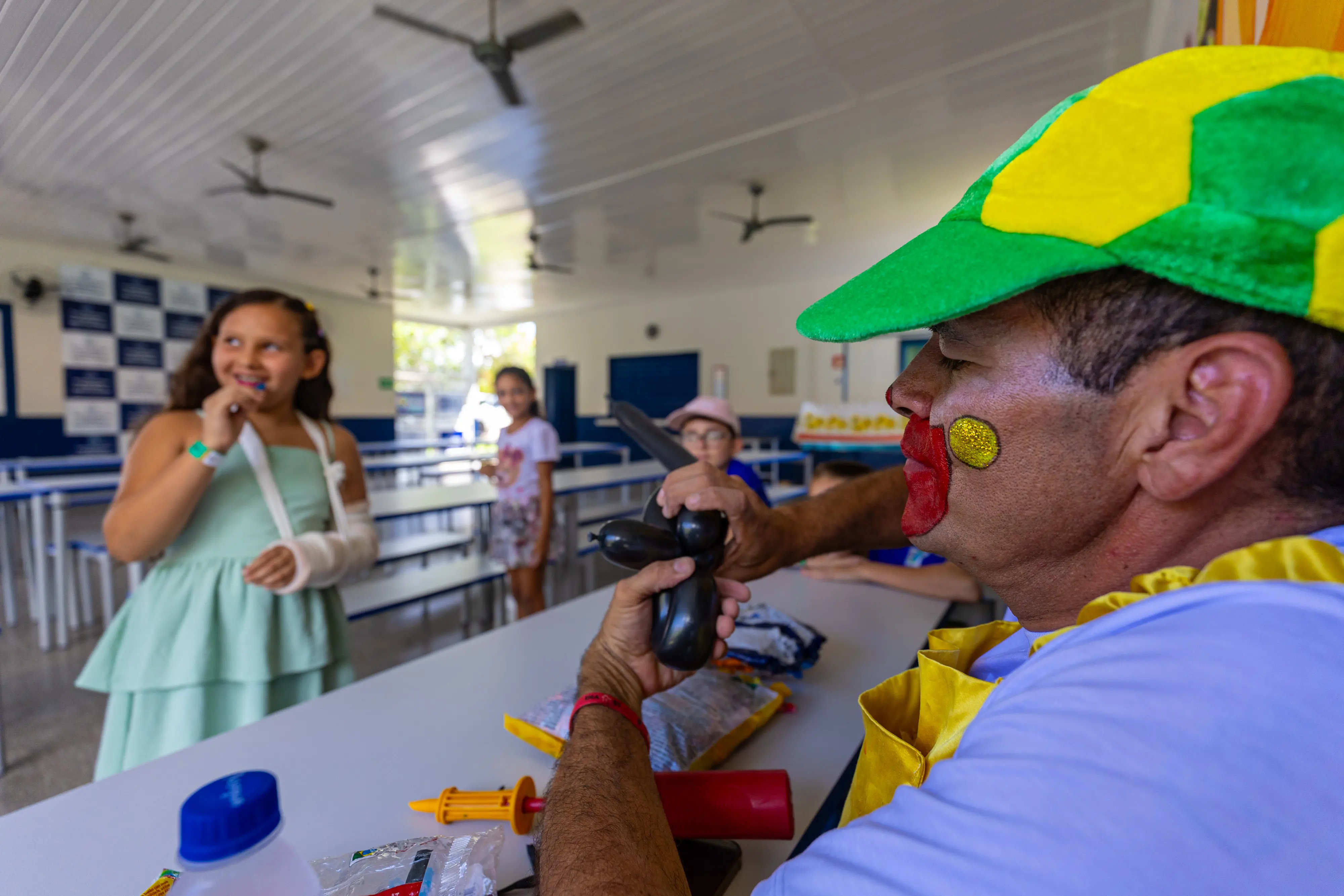 Um palhaço de perfil molda um balão preto para crianças em um refeitório. À esquerda, uma menina de vestido verde e braço imobilizado sorri. O palhaço usa chapéu verde e amarelo e pintura facial.