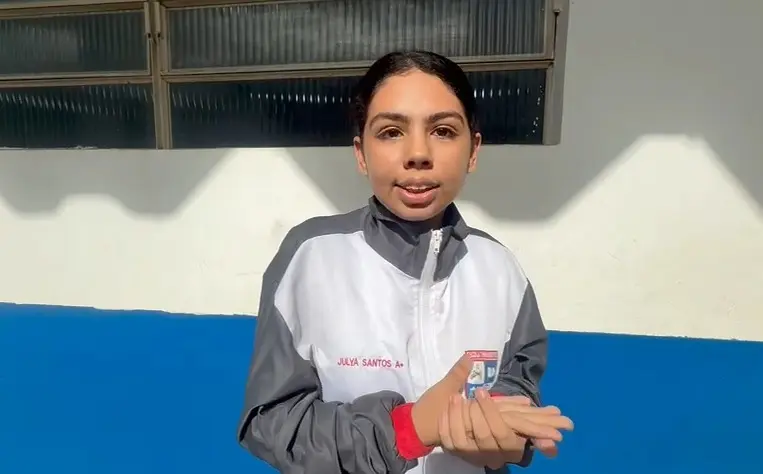 Foto horizontal em plano médio que mostra a aluna da Escola Tirandentes de Várzea Grande, Ana Júlya Tavares em pé, gesticulando. Ela é uma adolescente de pele clara, olhos castanhos, cabelo castanho preso, usando moletom cinza e branco da escola militar.