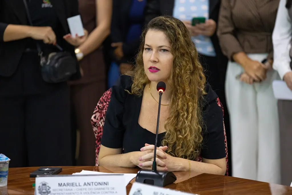 Foto horizontal que mostra a secretária-chefe do Gabinete de Enfrentamento à Violência Contra a Mulher, Mariell Antonini, falando ao microfone, sentada. Ela é uma mulher branca, de olhos azuis, longos cabelos loiros e cacheados, usando vestido preto e batom cor-de-rosa.