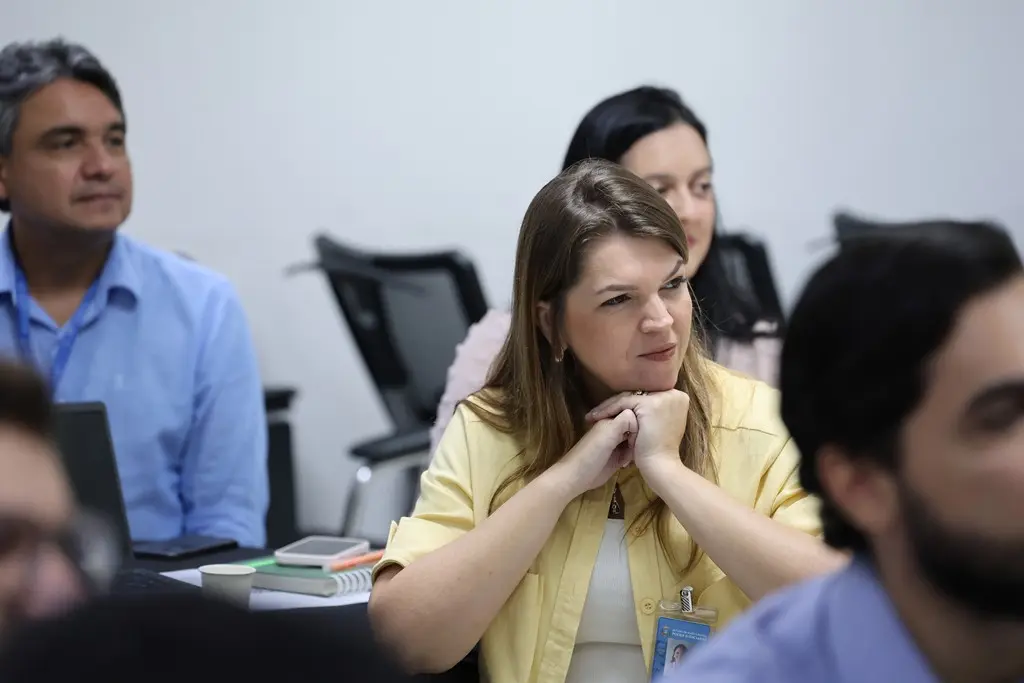Foto horizontal em plano médio, que mostra a servidora do TJMT, Jaqueline Schoffen, sentada em meio a outros participantes da oficina de inovação. ela é uma mulher branca, loira, usando blusa branca e camisa amarela, sorrindo e olhando em direção ao professor.