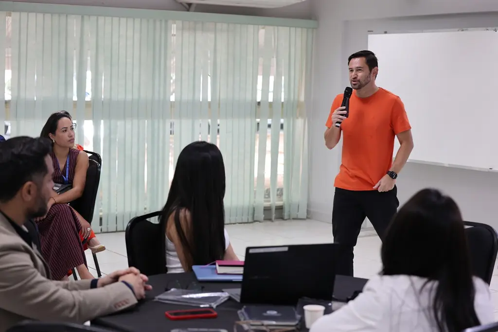 Foto horizontal em plano aberto que mostra uma sala de aula onde ocorre a oficina de inovação. O instrutor André Temura está em pé, falando ao microfone, e os participantes estão sentados em volta de mesas redondas.