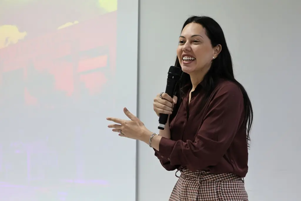 Foto horizontal em plano médio que mostra a juíza Joseane Antunes em pé, falando ao microfone, numa sala de aula. Ela é uma mulher branca, de cabelos longos e pretos, usando camisa marrom e sorrindo.