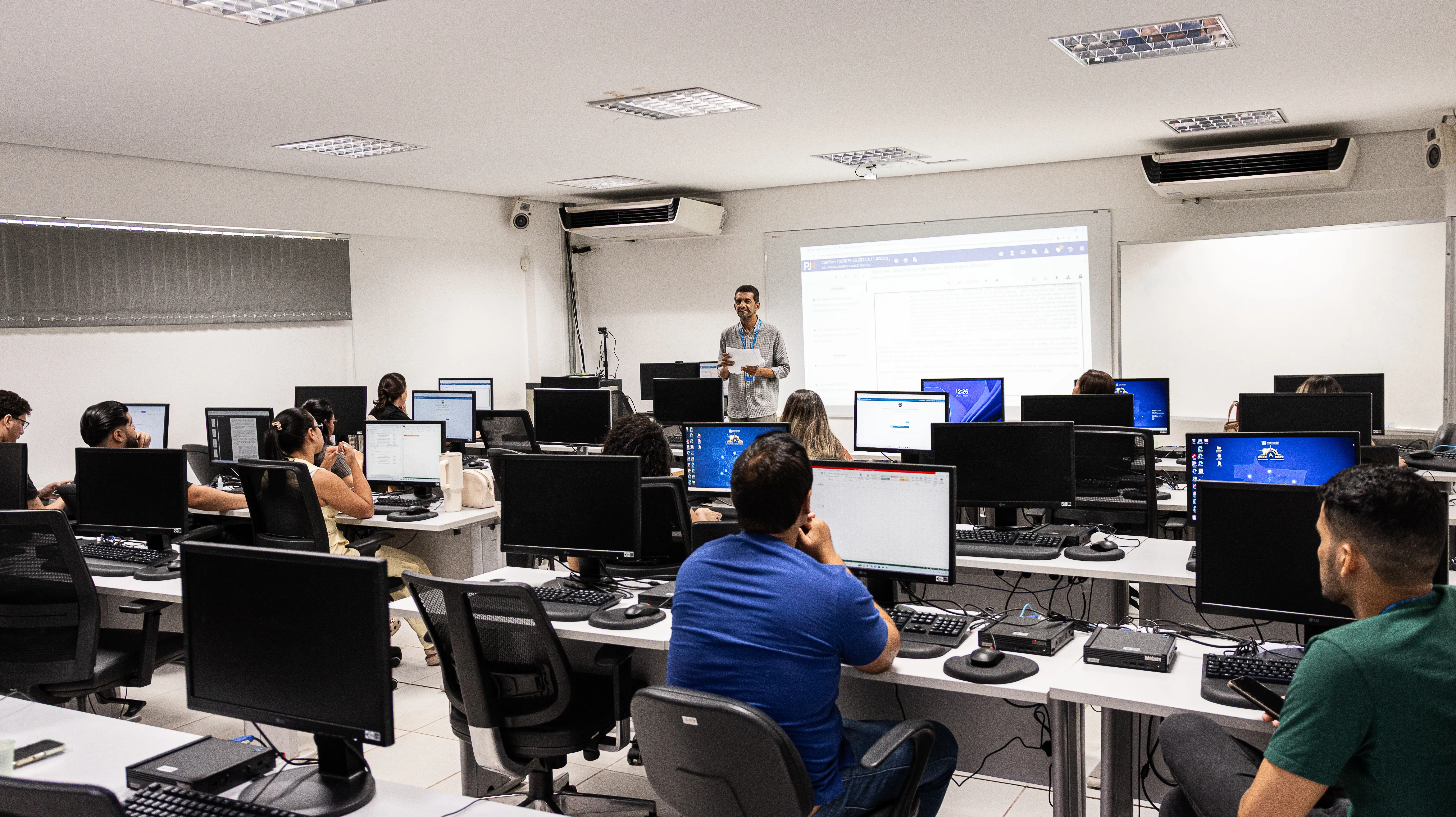 Instrutor em pé fala diante de uma sala de informática lotada, com alunos em bancadas usando computadores. Ao fundo, uma projeção exibe a tela de um sistema.