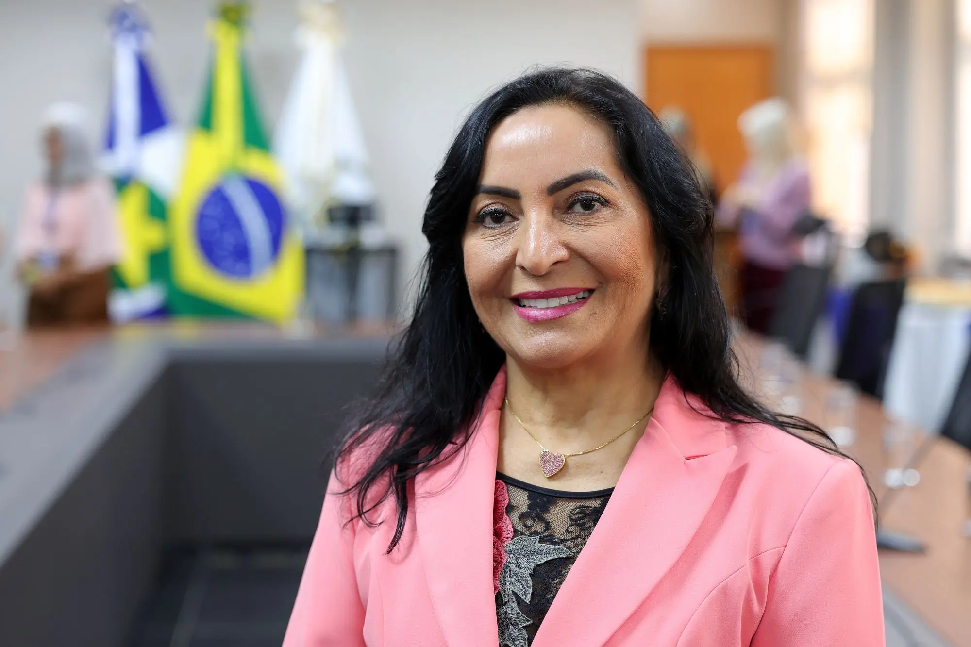 Foto horizontal que mostra Claudenice Deijany, do busto pra cima, sorrindo para a foto. Ela é uma mulher de meia idade, parda, de olhos escuros, longos cabelos pretos e lisos, usando blusa preta e blazer rosa. O fundo desfocado mostra uma sala de reuniões com pessoas e mastros com bandeiras.