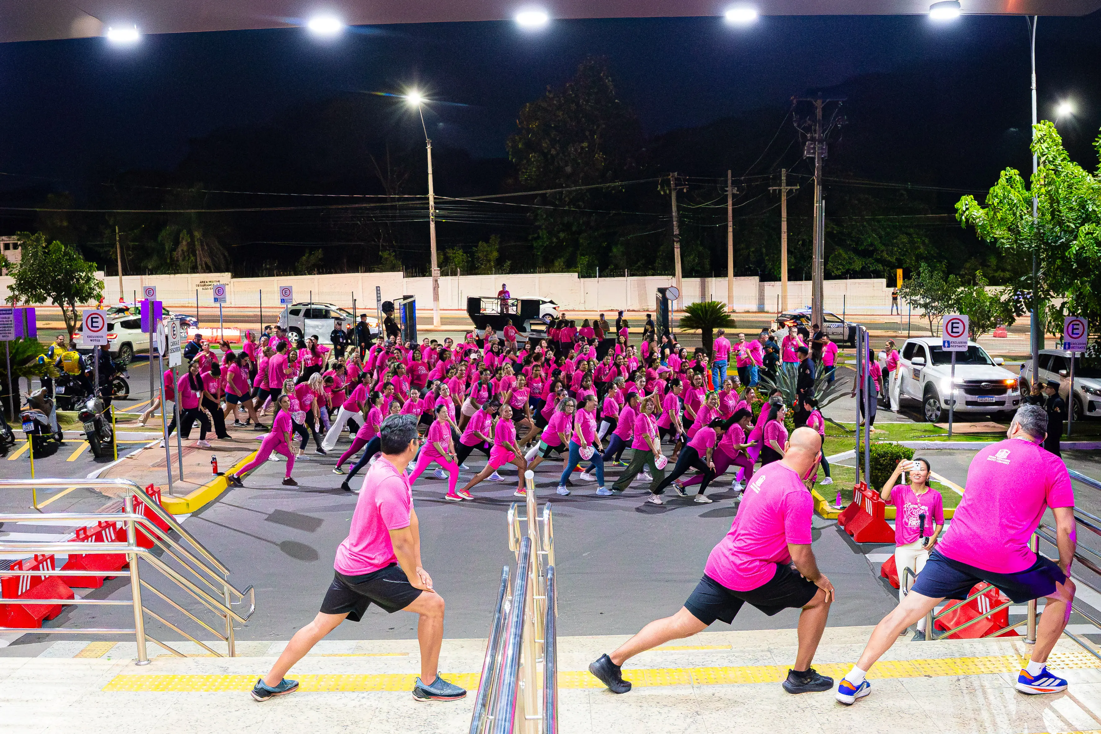 Grande grupo de pessoas com camisetas cor-de-rosa realiza exercícios de alongamento ao ar livre à noite. Em primeiro plano, três pessoas se preparam na escadaria.