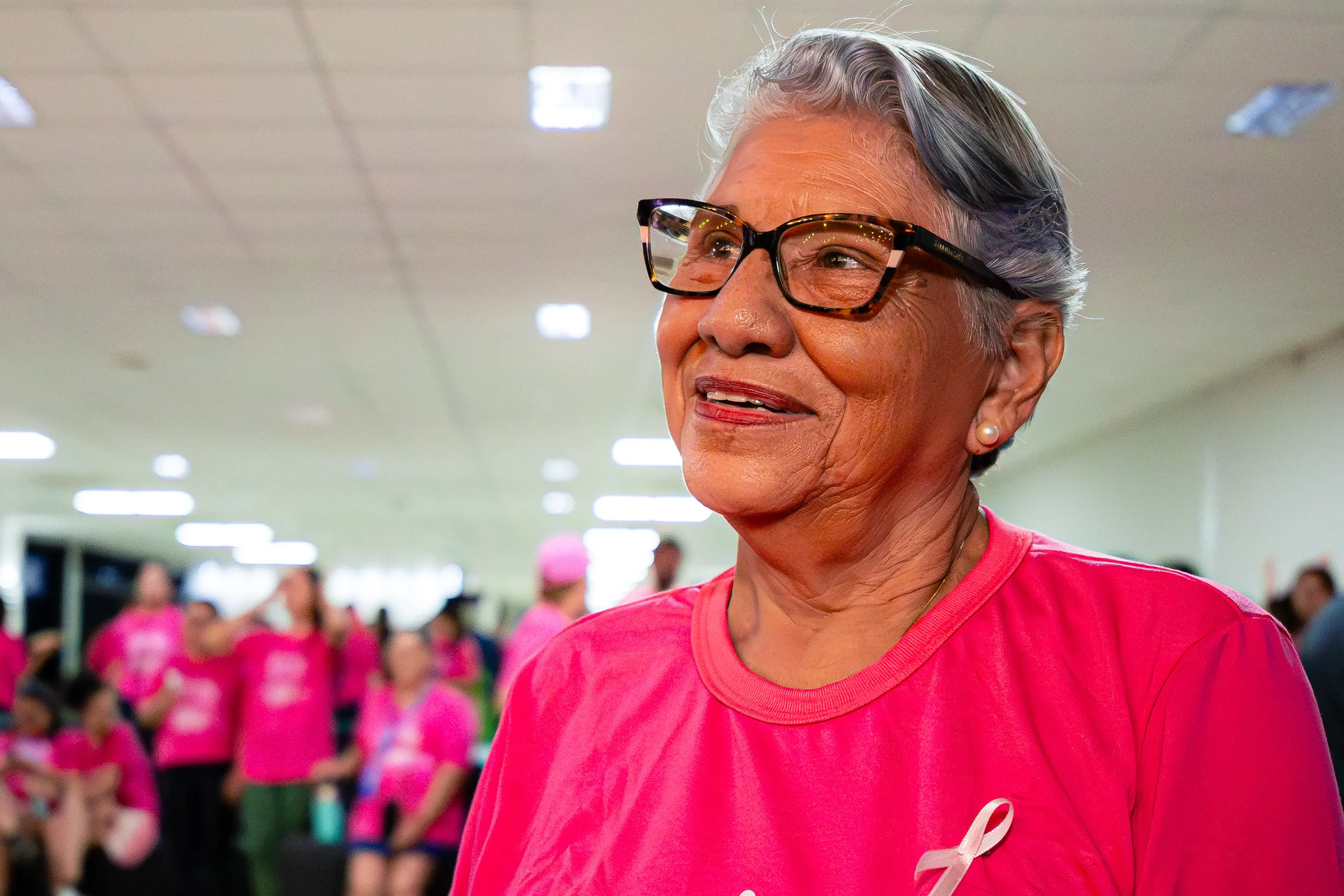 Mulher idosa sorri suavemente, usando óculos de armação escura e blusa rosa com um laço simbólico. Ao fundo, pessoas com vestimentas semelhantes em um ambiente interno iluminado.