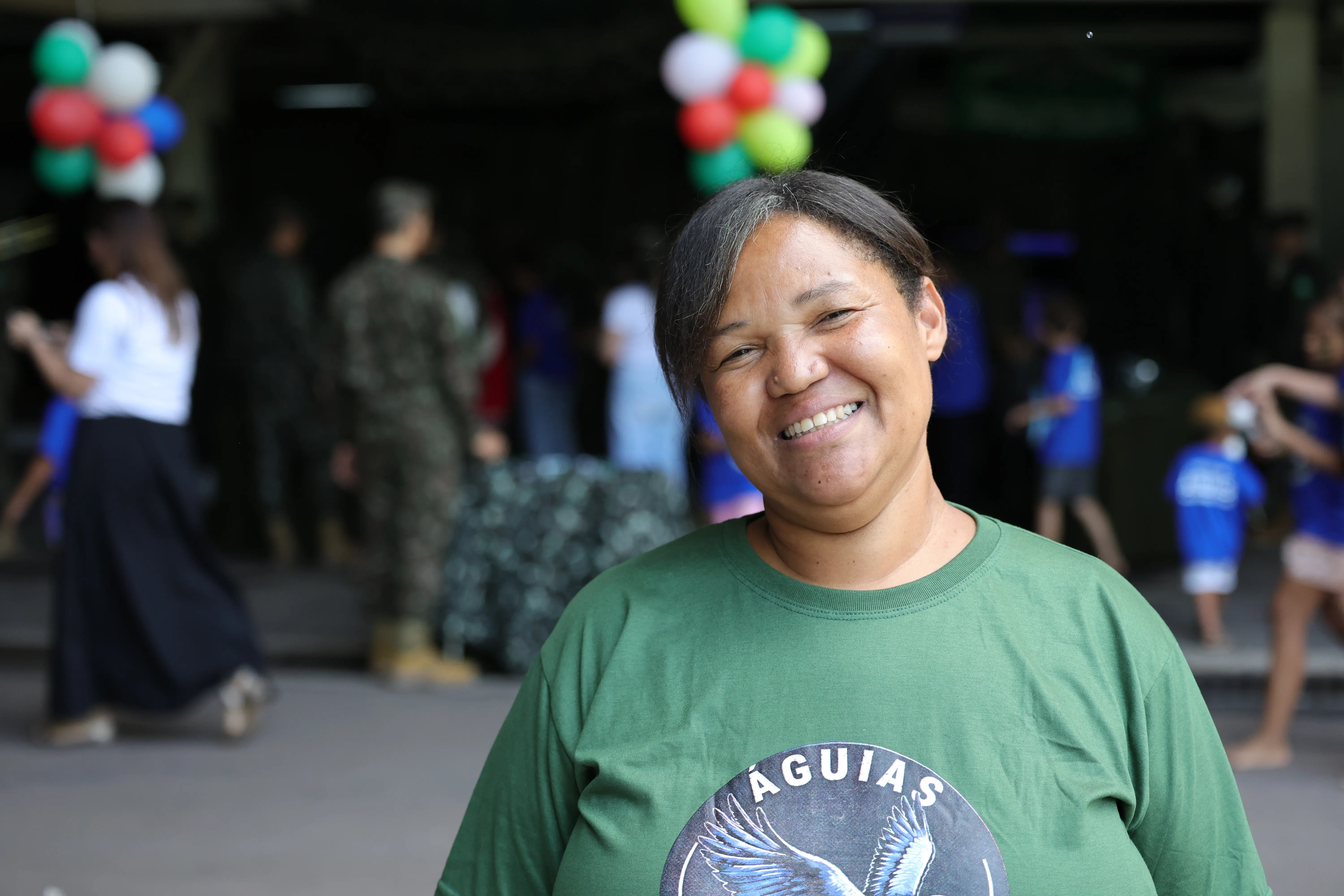 Mulher sorridente de camiseta verde com a palavra "ÁGUIAS" em destaque. Ao fundo desfocado, há pessoas movimentando-se e balões coloridos suspensos.