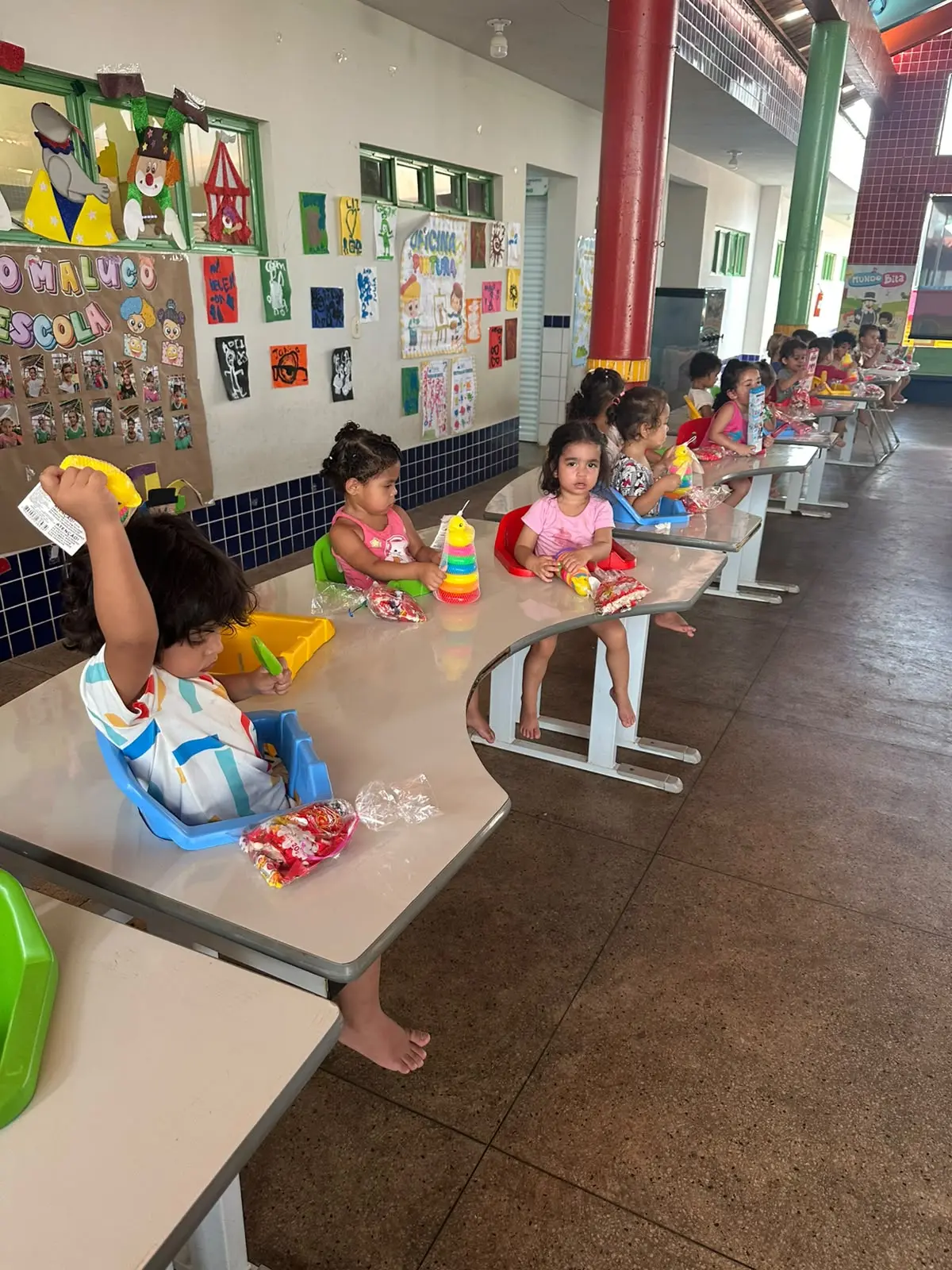 Crianças pequenas estão sentadas em cadeirinhas acopladas a mesas em fileira em um corredor escolar. Sobre as mesas há brinquedos coloridos e sacos de doces; as paredes têm desenhos infantis.