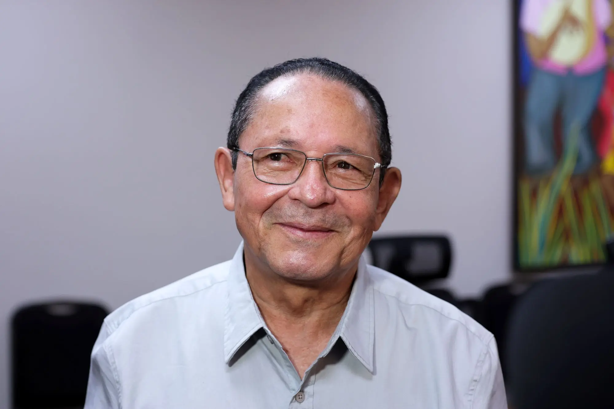 Foto horizontal que mostra o servidor aposentado Leônidas Palmeira, do busto pra cima, sorrindo. Ele é um senhor idoso, de cabelos e olhos escuros. Ele usa óculos de grau e camisa social cinza clara.