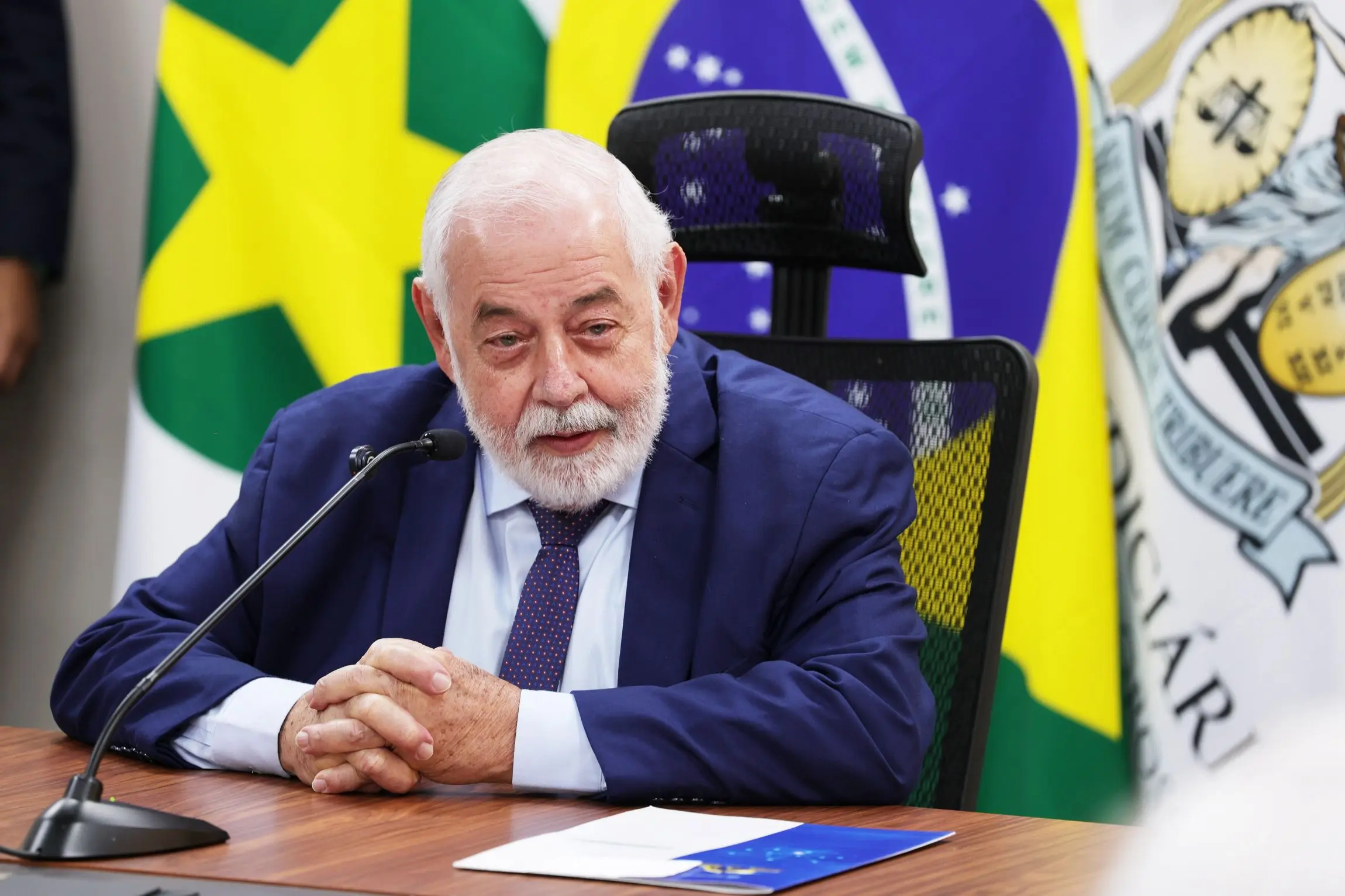 Foto horizontal que mostra o desembargador José Zuquim Nogueira sentado em uma poltrona, com os braços sobre uma mesa de madeira e falando ao microfone. Ele é um senhor branco, de cabelo e barba brancos, olhos azuis. ele usa camisa branca, terno e gravata azul marinho. Ao fundo há bandeiras.