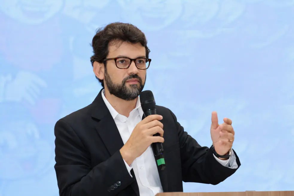 A imagem mostra uma pessoa em pé atrás de um púlpito ou mesa de apoio, segurando um microfone com uma das mãos e gesticulando com a outra, em uma situação que aparenta ser uma apresentação, palestra ou exposição oral.