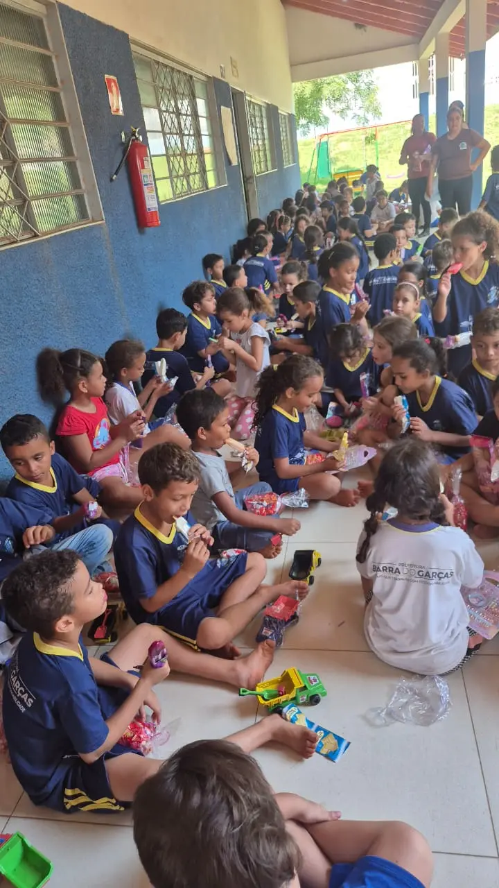 Crianças usando uniformes escolares azuis estão sentadas no chão ao longo de um corredor coberto. Elas manuseiam brinquedos e doces, com um extintor de incêndio visível na parede azul.