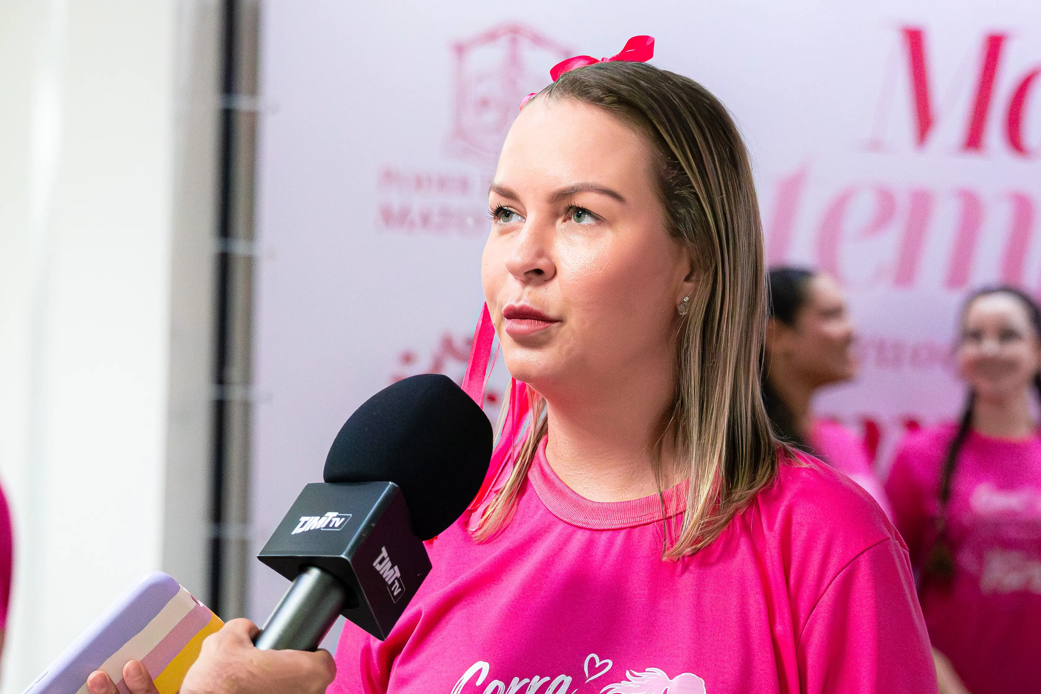 Mulher loira de blusa rosa fala ao microfone com a marca "TJMT TV". Ao fundo, outras pessoas vestindo rosa em ambiente interno.