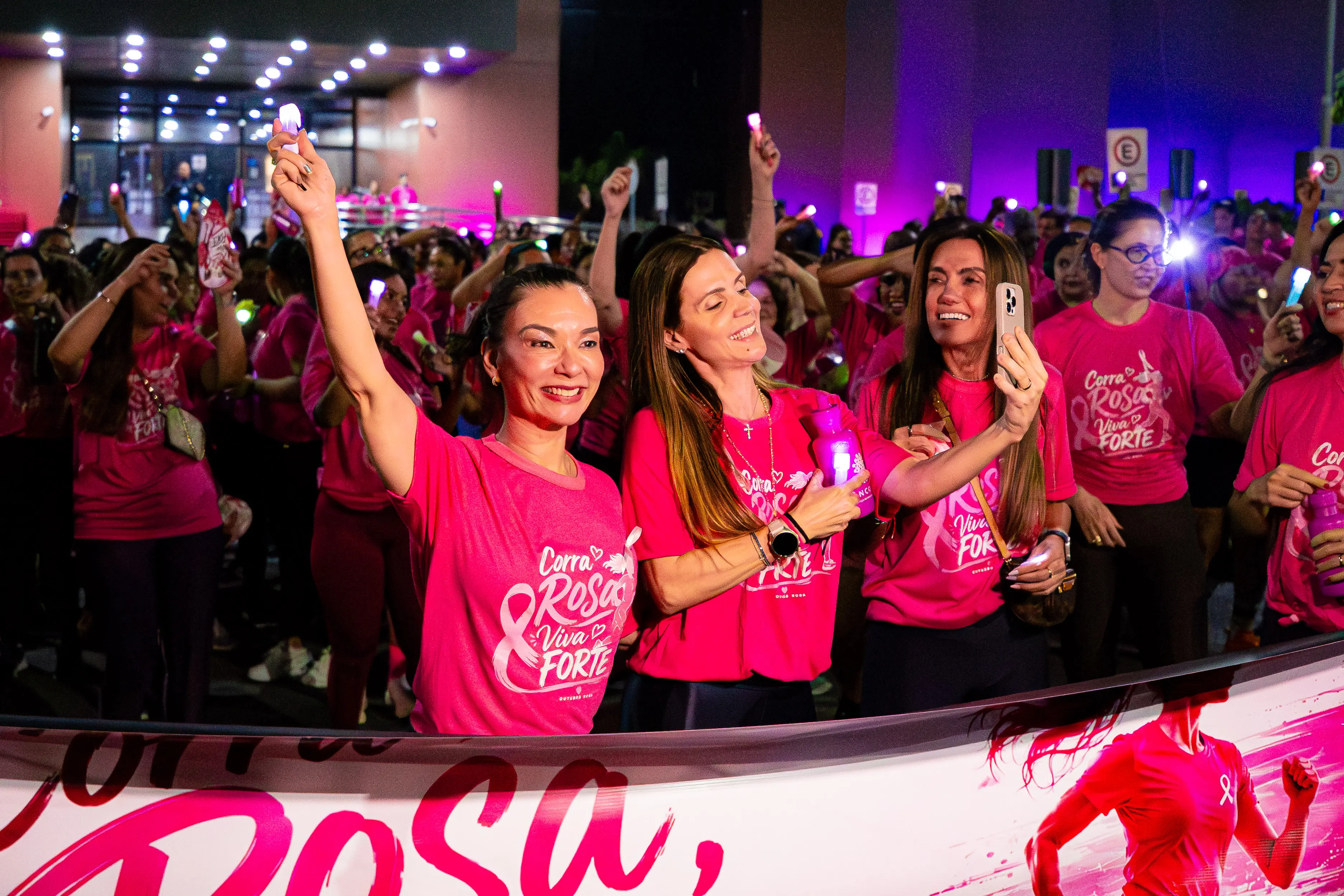 Mulheres com camisetas rosas estampadas participam de evento noturno com iluminação temática. Em primeiro plano, três mulheres posam sorridentes, segurando uma garrafa e um celular.