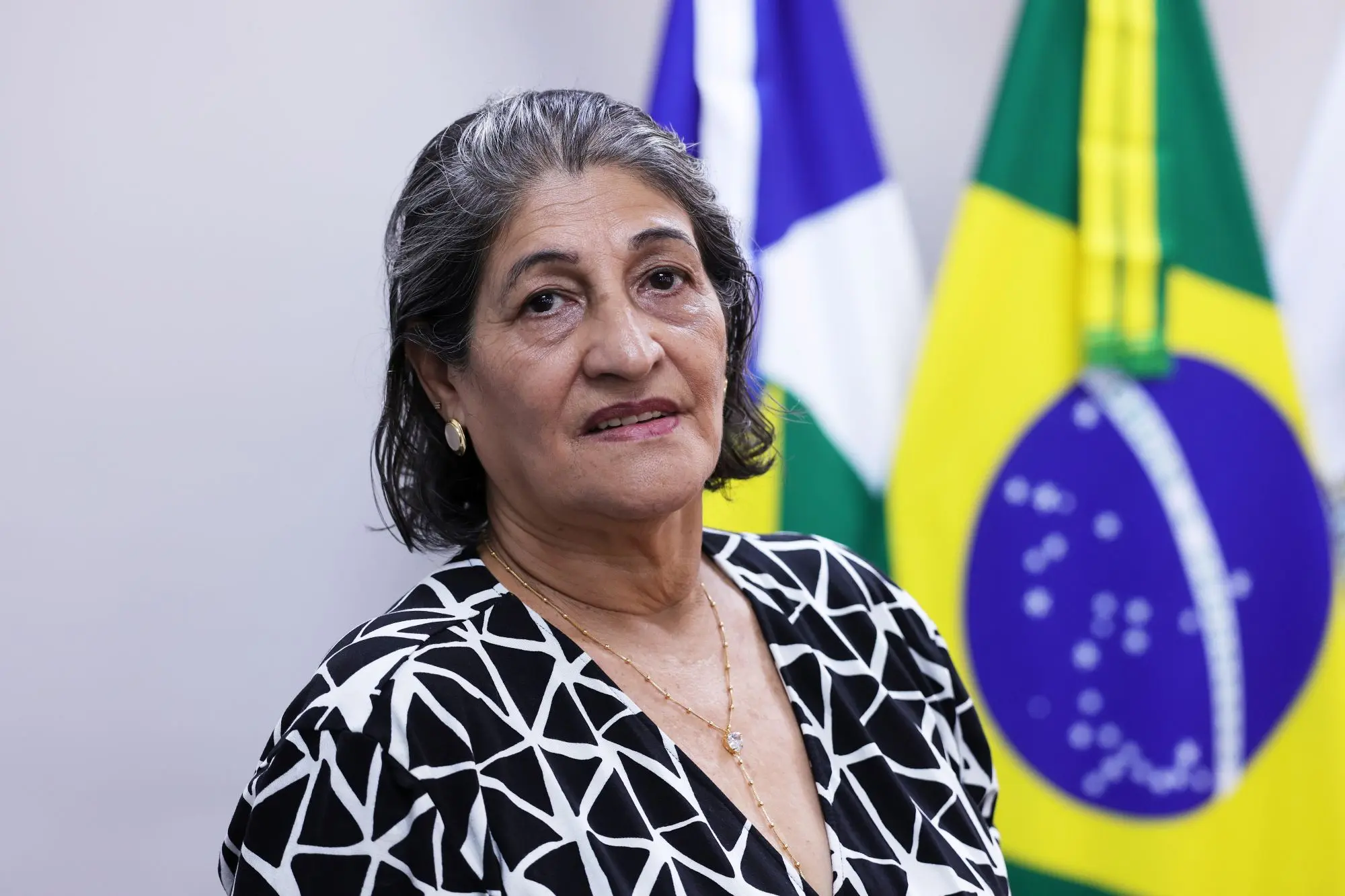 Foto horizontal que mostra a servidora aposentada Suely Rodrigues, do busto pra cima, olhando pra foto. Ela é uma senhora de pele clara, cabelos lisos e grisalhos, na altura dos ombros, usando vestido preto com estampa geométrica branca. Atrás dela aparecem bandeiras do Brasil e de Mato Grosso.