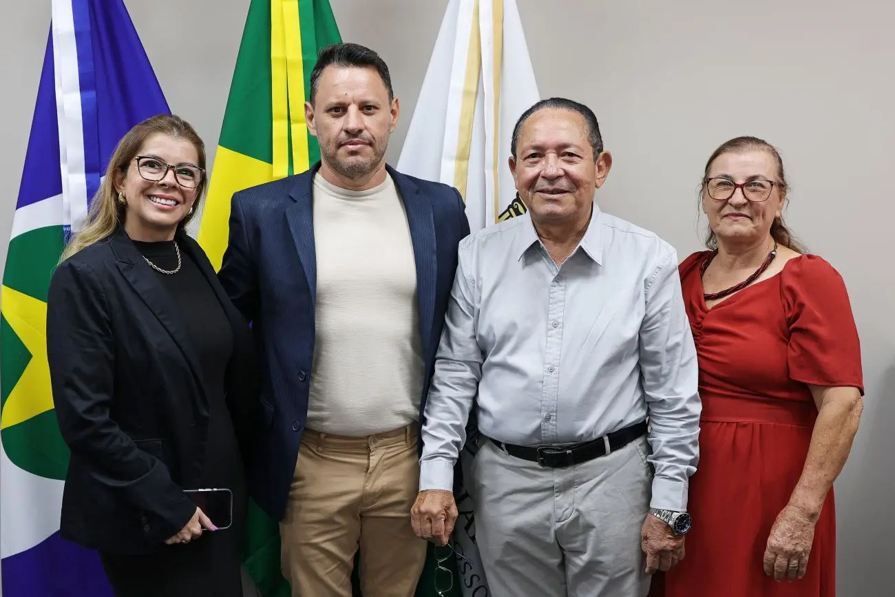 Foto horizontal que mostra o servidor aposentado Leônidas Palmeira, ladeado pela esposa, filho e nora, em pé, em frente a mastros com bandeiras. Eles sorriem para a foto.