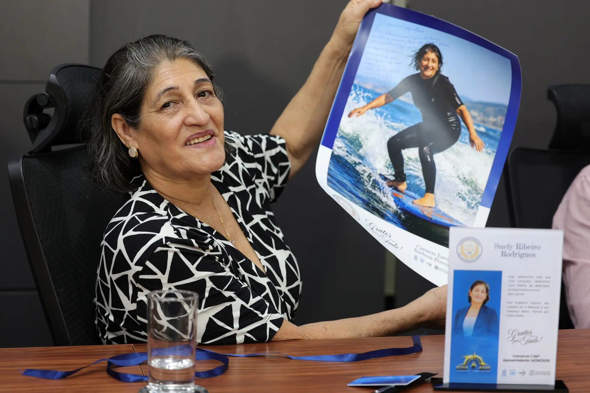 Foto horizontal que mostra a servidora aposentada Suely Rodrigues em uma sala de reuniões, sentada. Ela olha para a foto e mostra um pôster com uma imagem dela criada por inteligência artificial surfando. sobre a mesa há uma placa de homenagem com a foto dela.