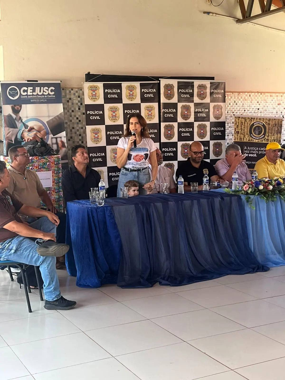 Uma mulher fala ao microfone no centro de uma mesa revestida por tecido azul, acompanhada por homens sentados. Ao fundo, há painéis com o logotipo da Polícia Civil e do CEJUSC.
