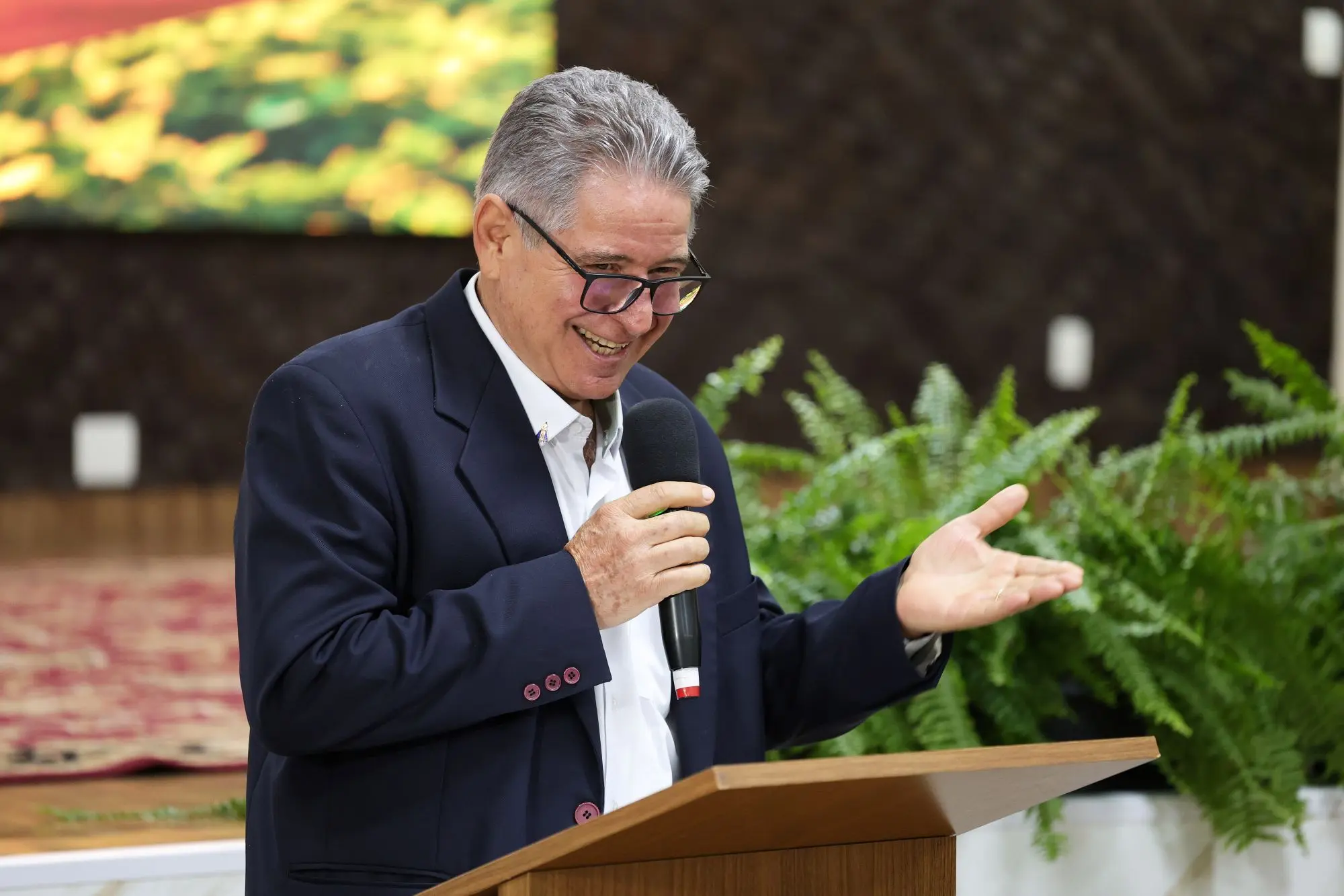 Foto horizontal que mostra o presidente da Famato, Vilmondes Tomain, atrás de um púlpito de madeira, falando ao microfone, gesticulando e sorrindo, no auditório do TJMT. Ele é um senhor branco, de cabelos grisalhos, usando óculos de grau, camisa branca e paletó preto.