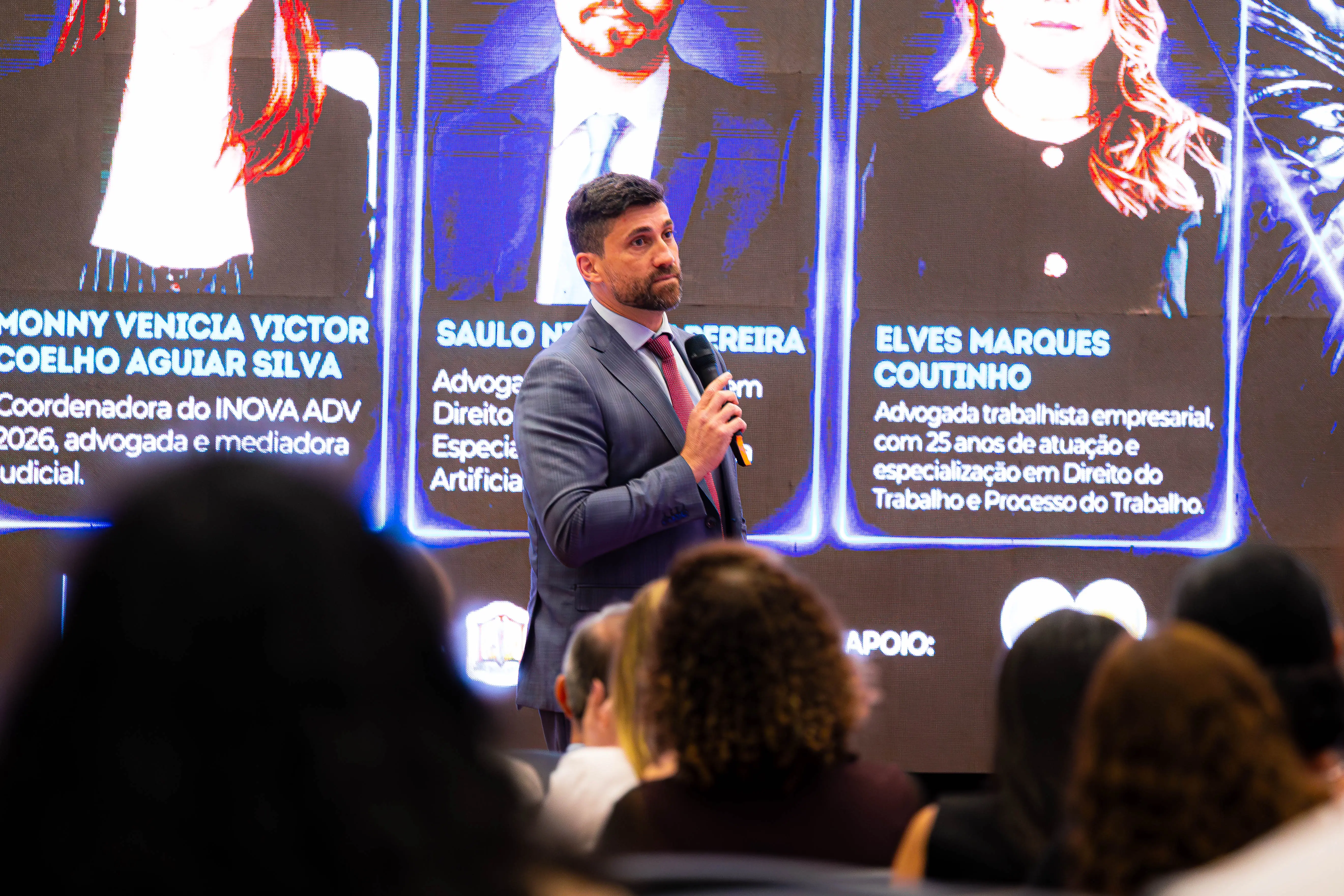 Homem de terno cinza e gravata vermelha fala ao microfone em pé diante da plateia. Ao fundo, um painel digital exibe fotos e textos informativos.