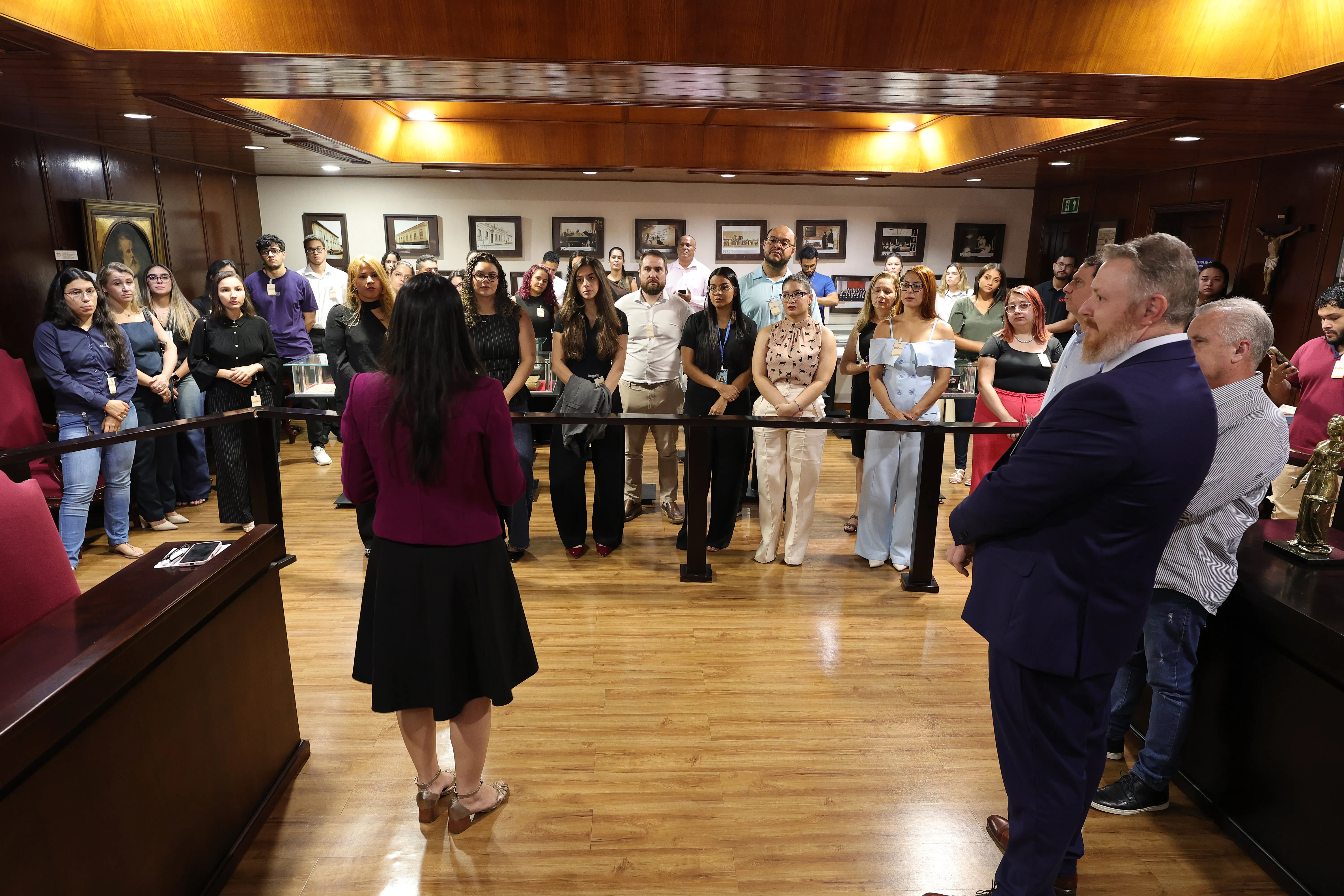 Mulher de saia preta e blazer roxo, vista de costas, fala para um grupo de pessoas em pé em plenário com acabamento em madeira. À direita, homens de terno observam a cena.