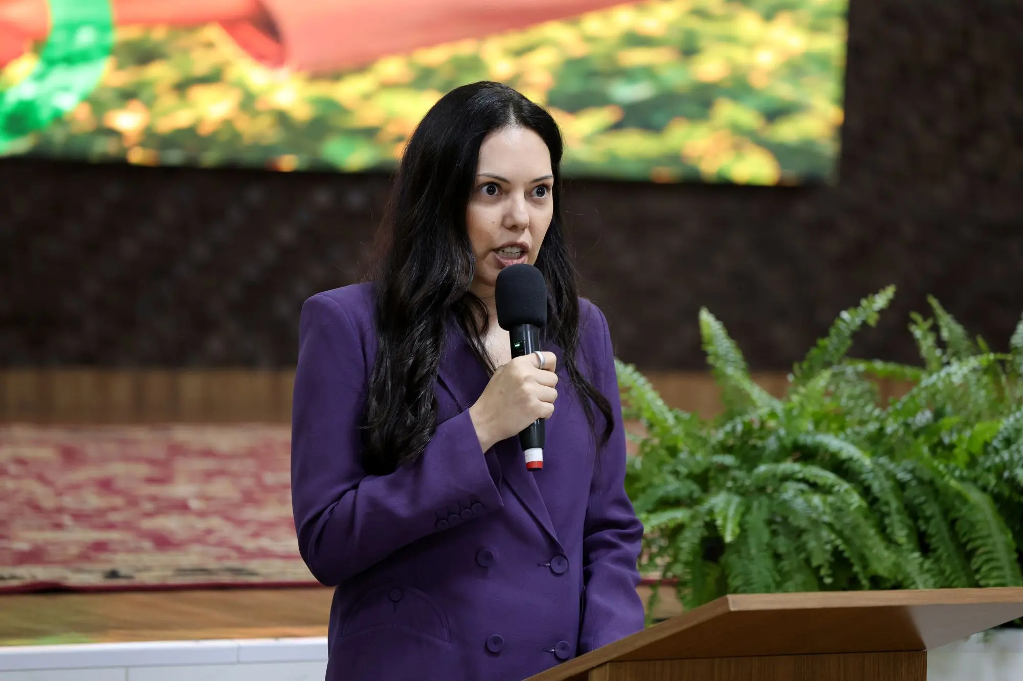 Foto horizontal que mostra a juíza coordenadora do Cejusc do Agro, Joseane Antunes, falando ao microfone, atrás do púlpito do auditório. Ela é uma mulher jovem, branca, de olhos pretos, cabelos pretos, longos e lisos, usando blazer roxo.