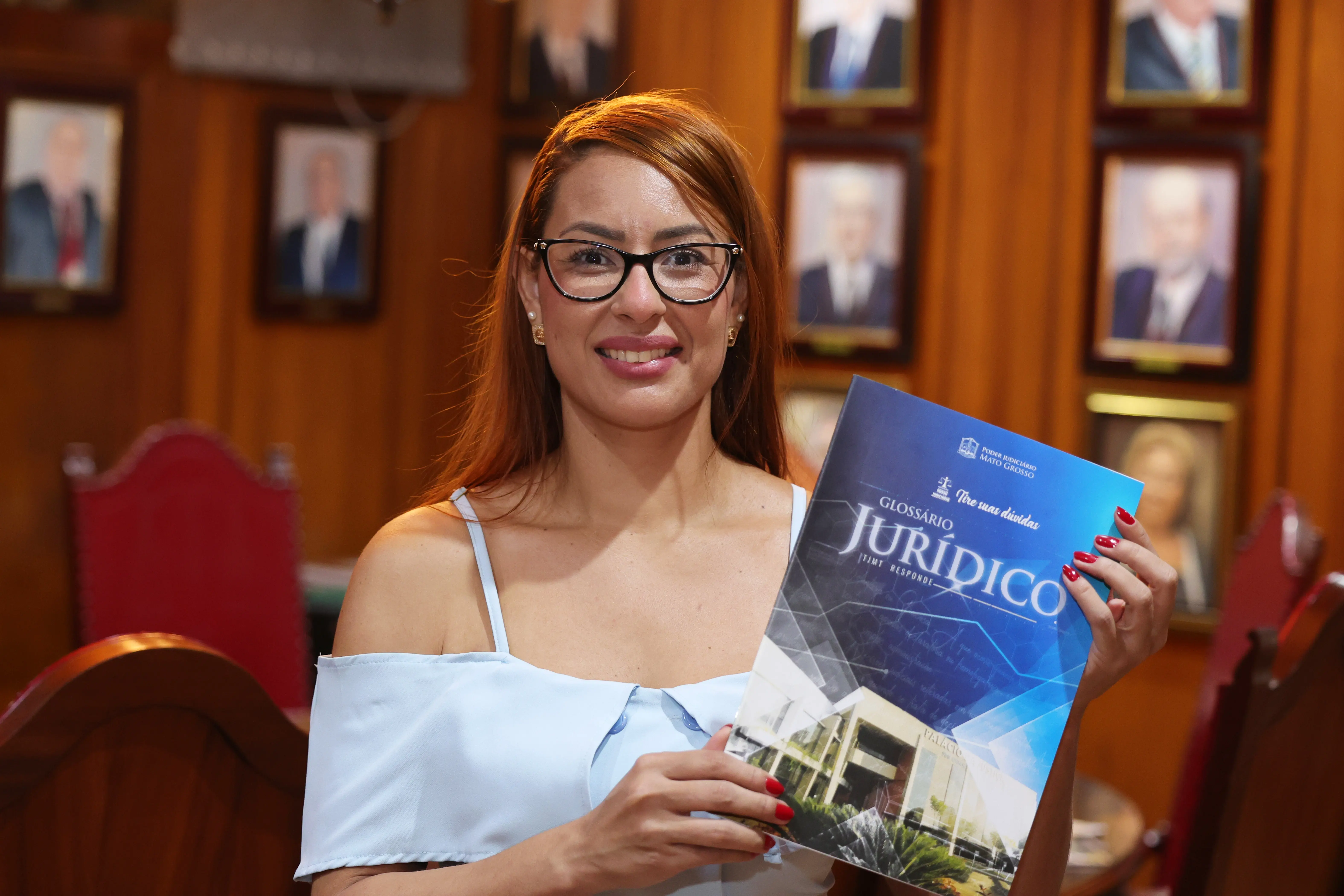 Mulher sorridente de óculos segura cartilha do "GLOSSÁRIO JURÍDICO". Usa blusa azul clara de ombros de fora. Ao fundo, parede de madeira com quadros de retratos.