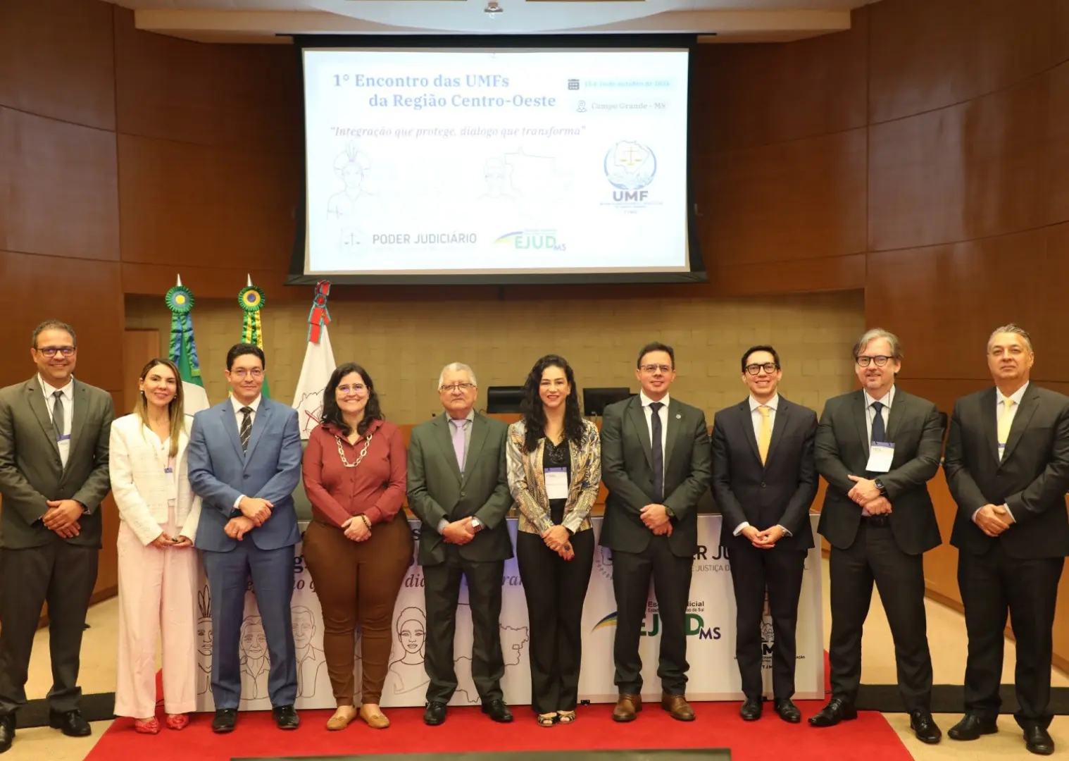 Foto em auditório com nove pessoas alinhadas lado a lado sobre um tapete vermelho. Elas vestem trajes formais como ternos, paletós e blusas sociais. Ao fundo, há telas com a projeção do "1º Encontro das UMFs da Região Centro-Oeste" e bandeiras oficiais.