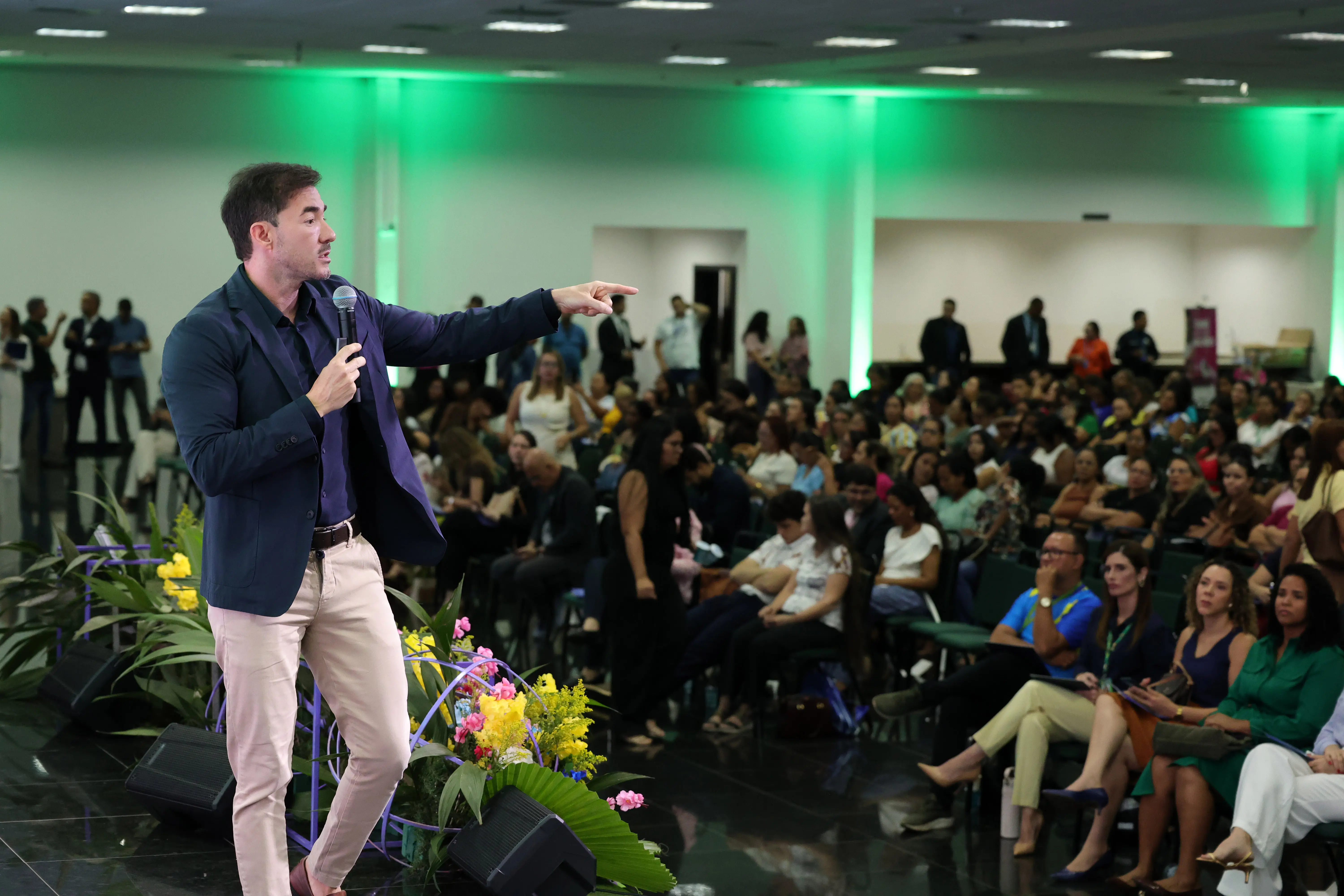 Homem de terno azul-marinho sem gravata e calça bege está em um auditório, apontando o dedo indicador direito para o público. Ele segura um microfone na outra mão. À frente há arranjos de flores e caixas de som, e uma plateia sentada assiste à palestra sob iluminação verde.