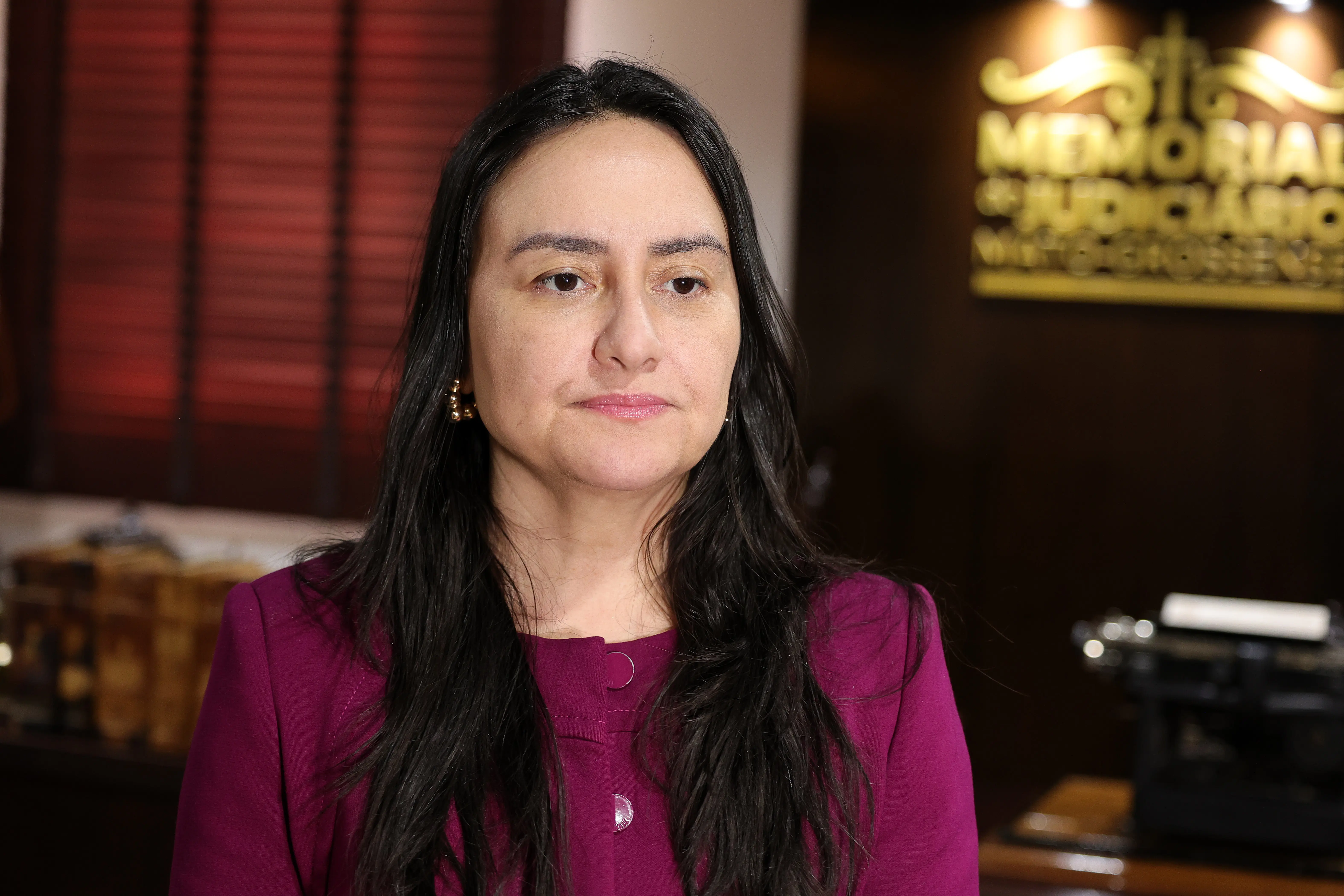 Mulher de cabelos longos e escuros veste blusa ou blazer roxo. Ao fundo, à direita, sobressai-se uma parede escura com a inscrição em letras douradas "MEMORIAL JUDICIÁRIO".