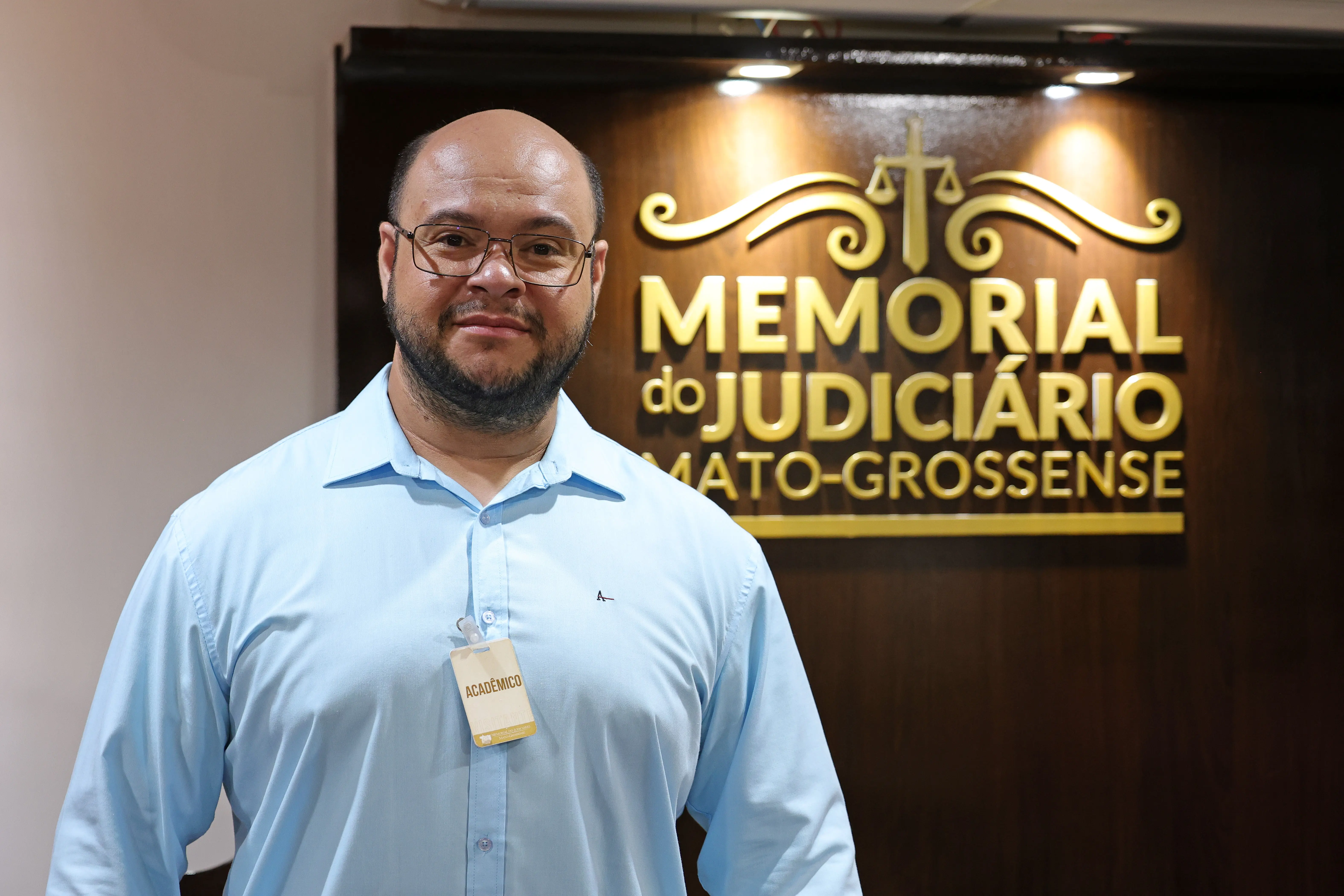 Homem de óculos e barba sorri vestindo camisa azul clara com crachá "ACADÊMICO". Ao fundo, a inscrição dourada "MEMORIAL do JUDICIÁRIO MATO-GROSSENSE".