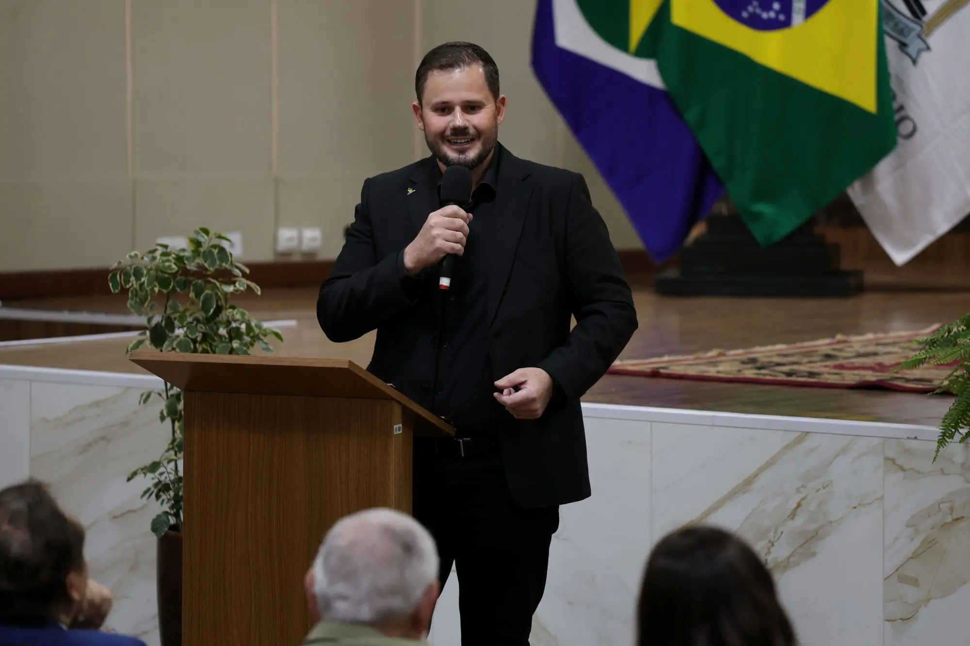 Foto horizontal que mostra o presidente da Aprosoja-MT, Lucas Costa Beber, falando ao microfone, no púlpito do auditório. Ele é um homem jovem, branco, de cabelos castanho claro, usando calça, camisa e paletó pretos.