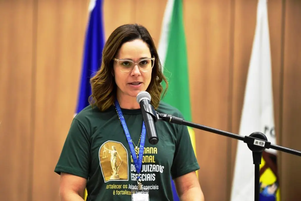 Na imagem a diretora do Daje, Shusiene Machado, fala ao microfone no púlpito. Ela é uma mulher branca, de cabelos castanhos, que usa óculos transparentes e está vestida com uma camisa verde que traz a logo da II Semana dos Juizados Especiais. Ao fundo estão três bandeiras incluindo a do Brasil