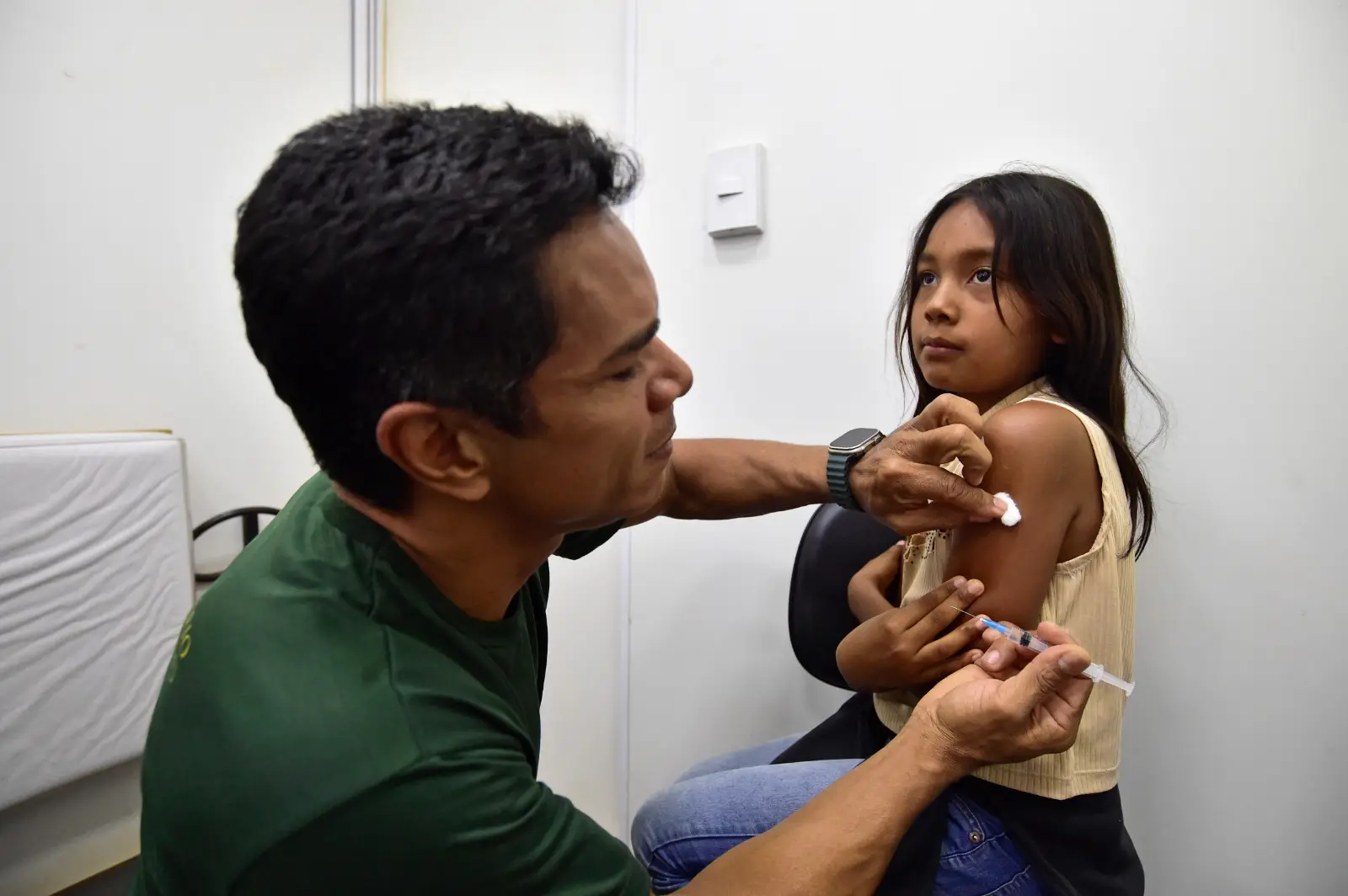 Homem aplica vacina no braço de uma menina com expressão serena. Eles estão em uma sala branca, com cadeira e cama ao fundo.