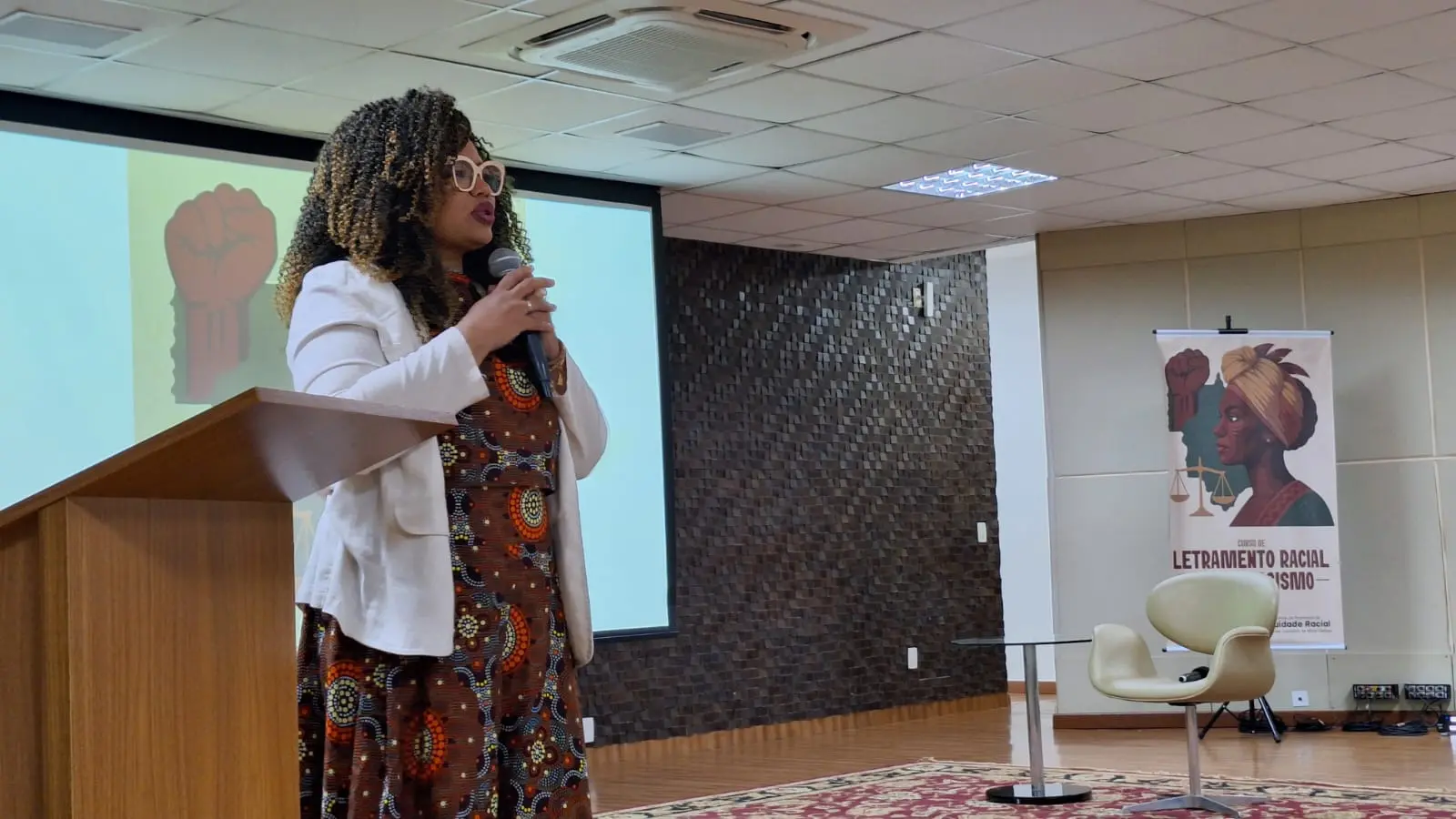Professora do curso de Letramento Racial, Silviane Ramos, fala ao microfone, em pé, no palco do auditório. Ela é uma mulher negra, com cabelos cacheados e com mechas douradas, usando vestido com estampa afro, blazer de linho branco e óculos de grau de armação bege. 
