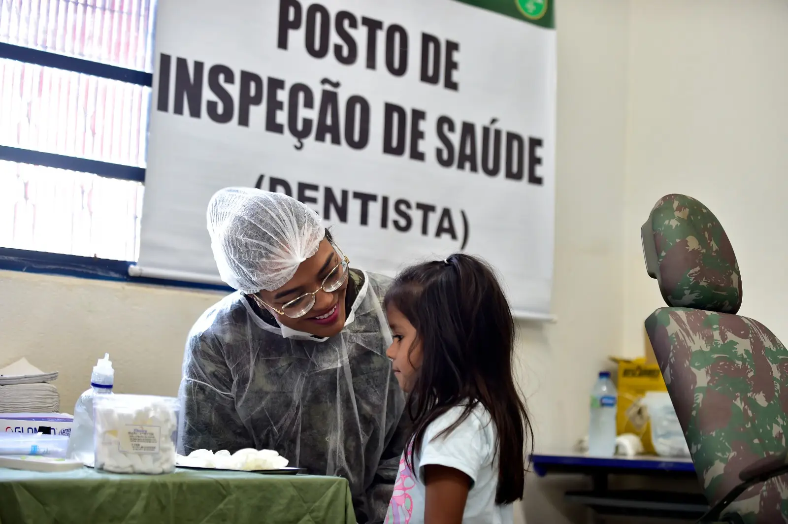 Dentista fardada, com touca e jaleco, sorri e conversa com menina em consultório improvisado. Ao fundo, placa de identificação do serviço odontológico.