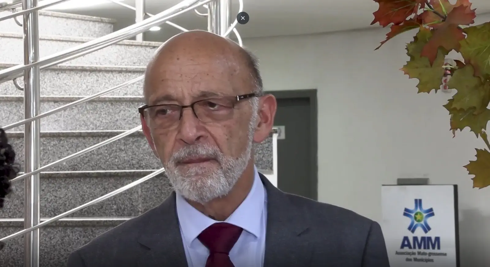 Alexandre Kalache Um homem mais velho, careca, com barba e óculos, veste um terno cinza e gravata bordô. Ele tem uma expressão séria e está em um ambiente interno com uma escada ao fundo e um logo da "AMM" visível.