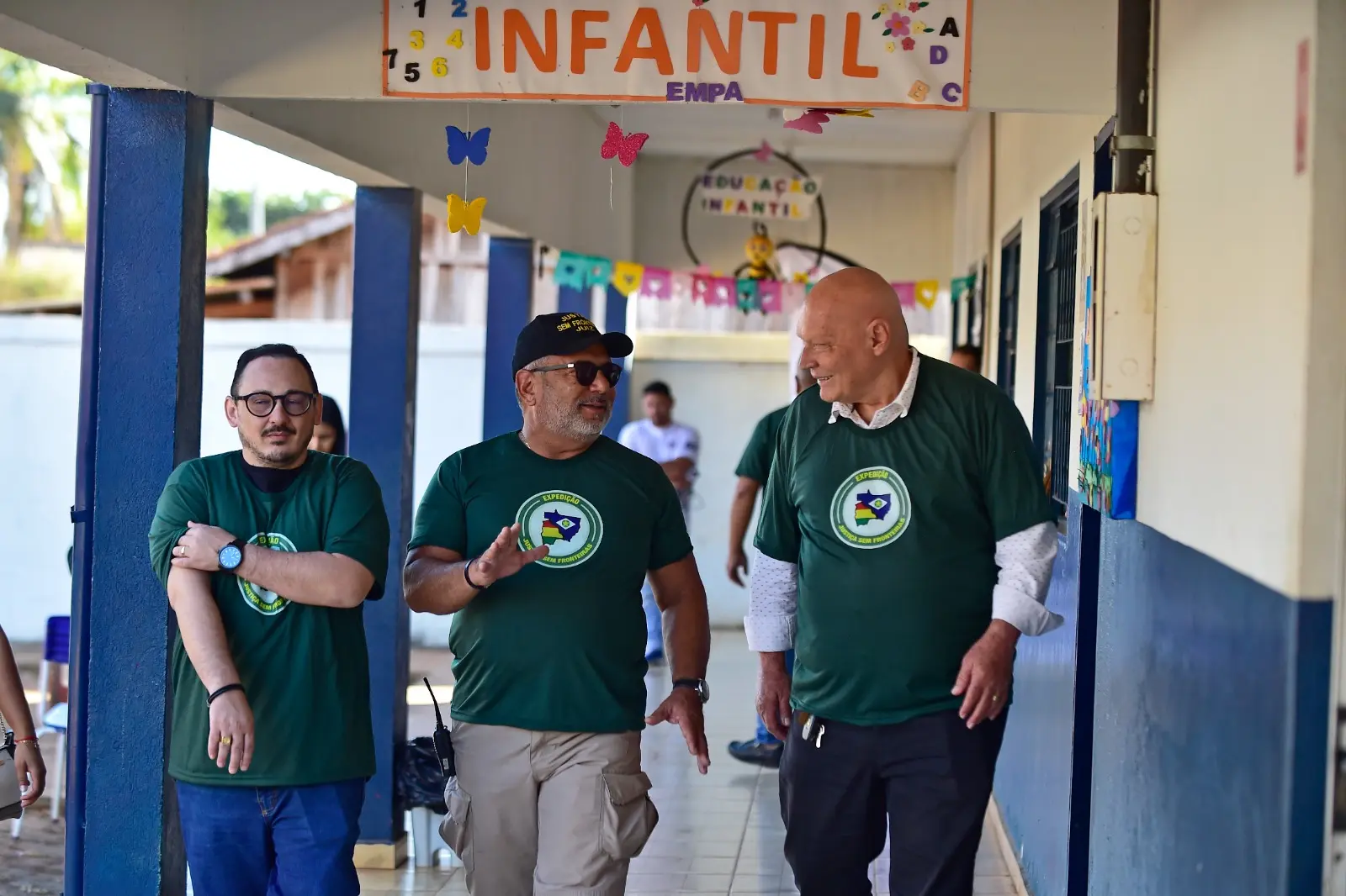 Três homens caminham sorrindo por corredor de escola com decoração infantil. Todos vestem camisetas verdes da Expedição Justiça Sem Fronteiras.