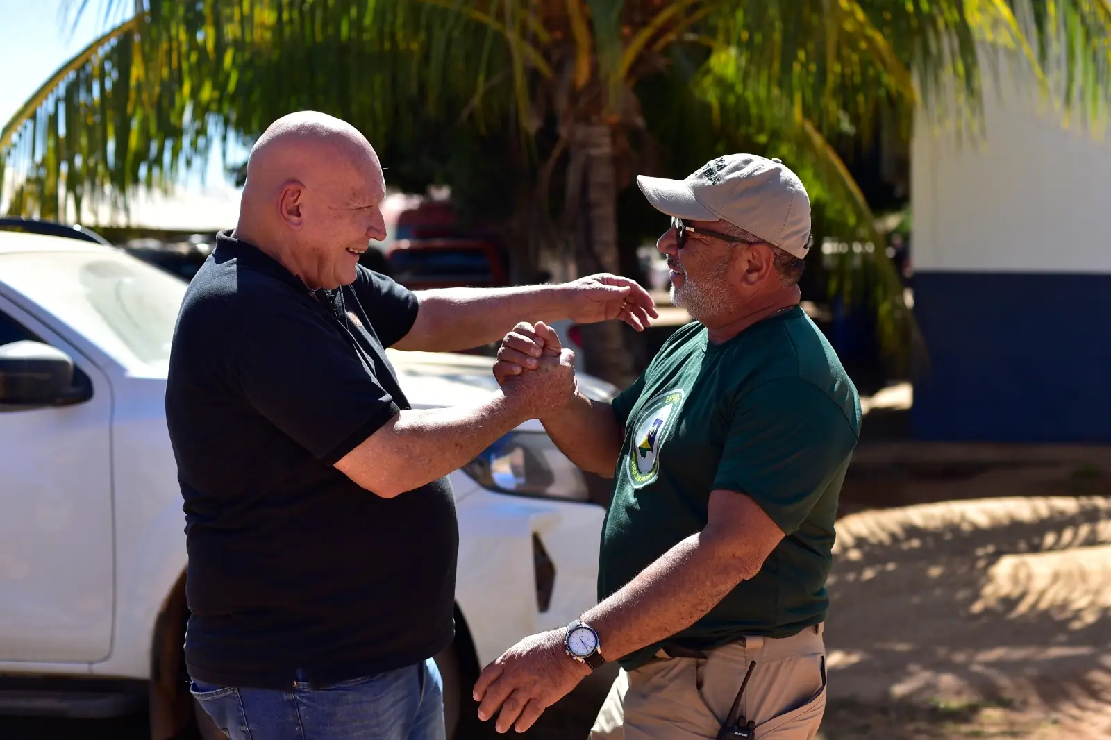 Dois homens se cumprimentam com alegria ao ar livre, trocando um aperto de mão afetuoso. Ao fundo, há um carro branco e folhas de palmeira sob a luz do sol.