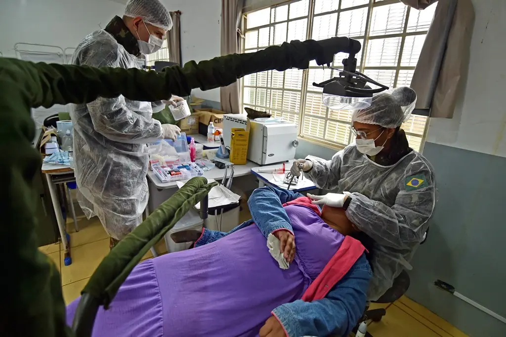 Foto que mostra uma mulher deitada na cadeira odontológica sendo atendida pela dentista e pelo técnico de saúde bucal do Exército. Ambos usam roupas militares e paramentos como capote, luva e touca.