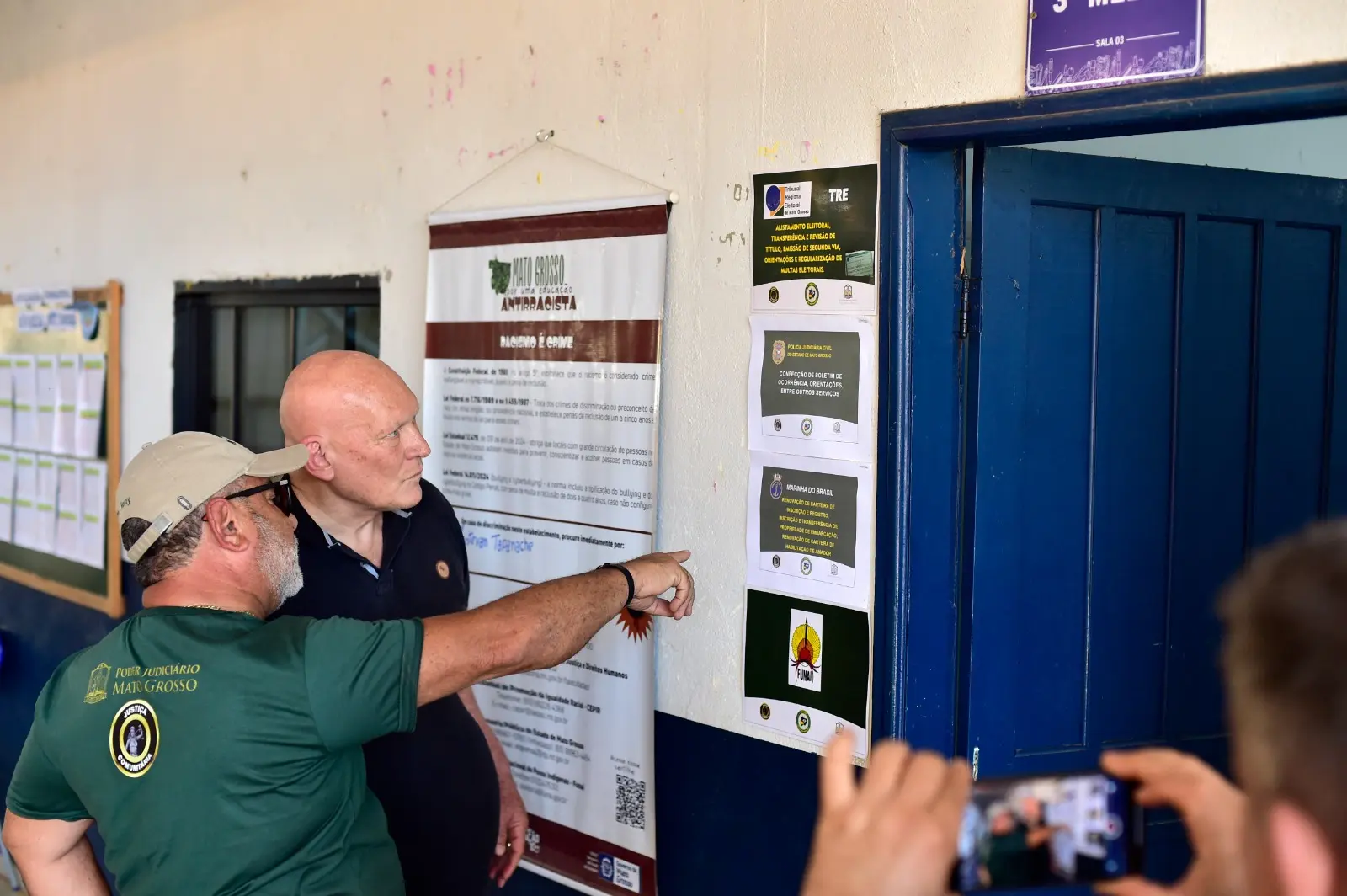 Dois homens observam e apontam para cartazes de orientação colados em parede escolar. Um deles usa camiseta do Poder Judiciário de Mato Grosso.