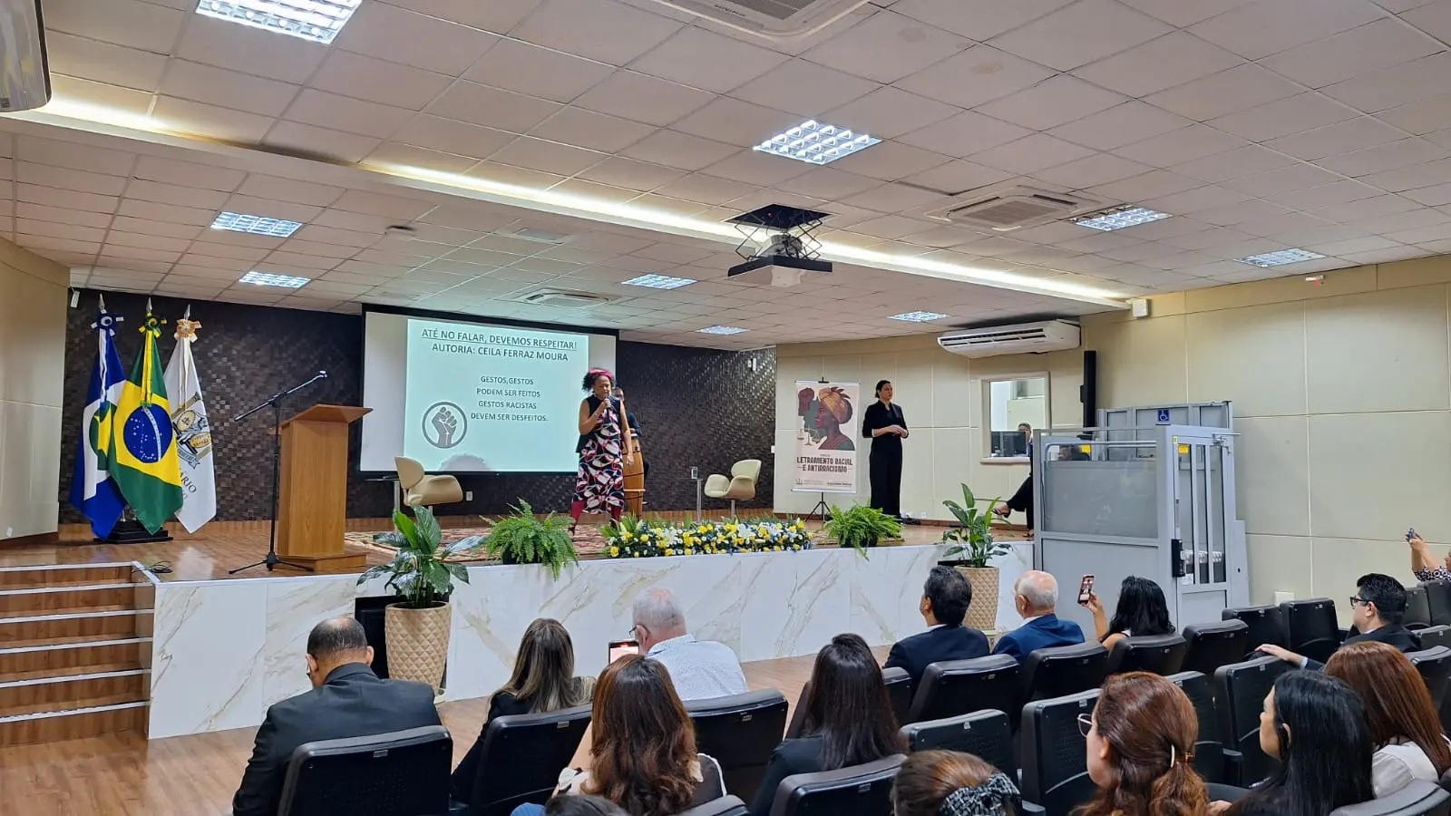 Foto em plano aberto que mostra a apresentação artística de Ceila Mônica e Regis Gomes. Ela está no centro do palco, com o microfone na mão. Ele está logo atrás, tocando atabaque. Há uma intérprete de Libras no palco. O público assiste sentado. 
