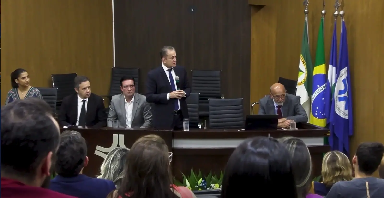 Uma visão frontal de um palco onde o Des. Orlando Perri de terno está de pé, falando ao centro, com outras quatro pessoas sentadas em uma mesa à frente. À direita, há três bandeiras. Várias pessoas no auditório assistem ao evento