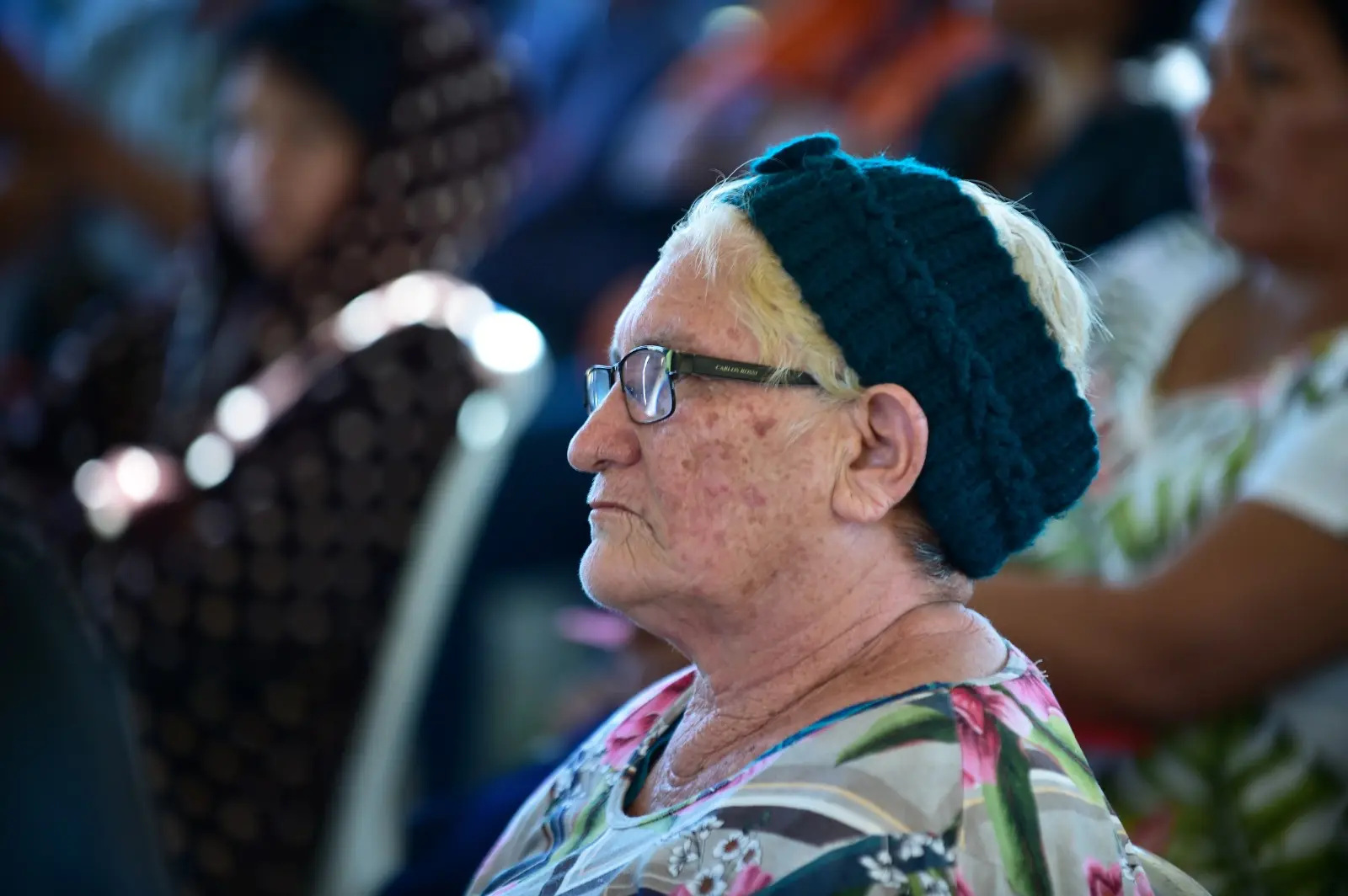 Idosa com cabelos claros, tiara azul e óculos observa a atividade ao redor. Ela veste roupa florida e está entre outros participantes do evento.
