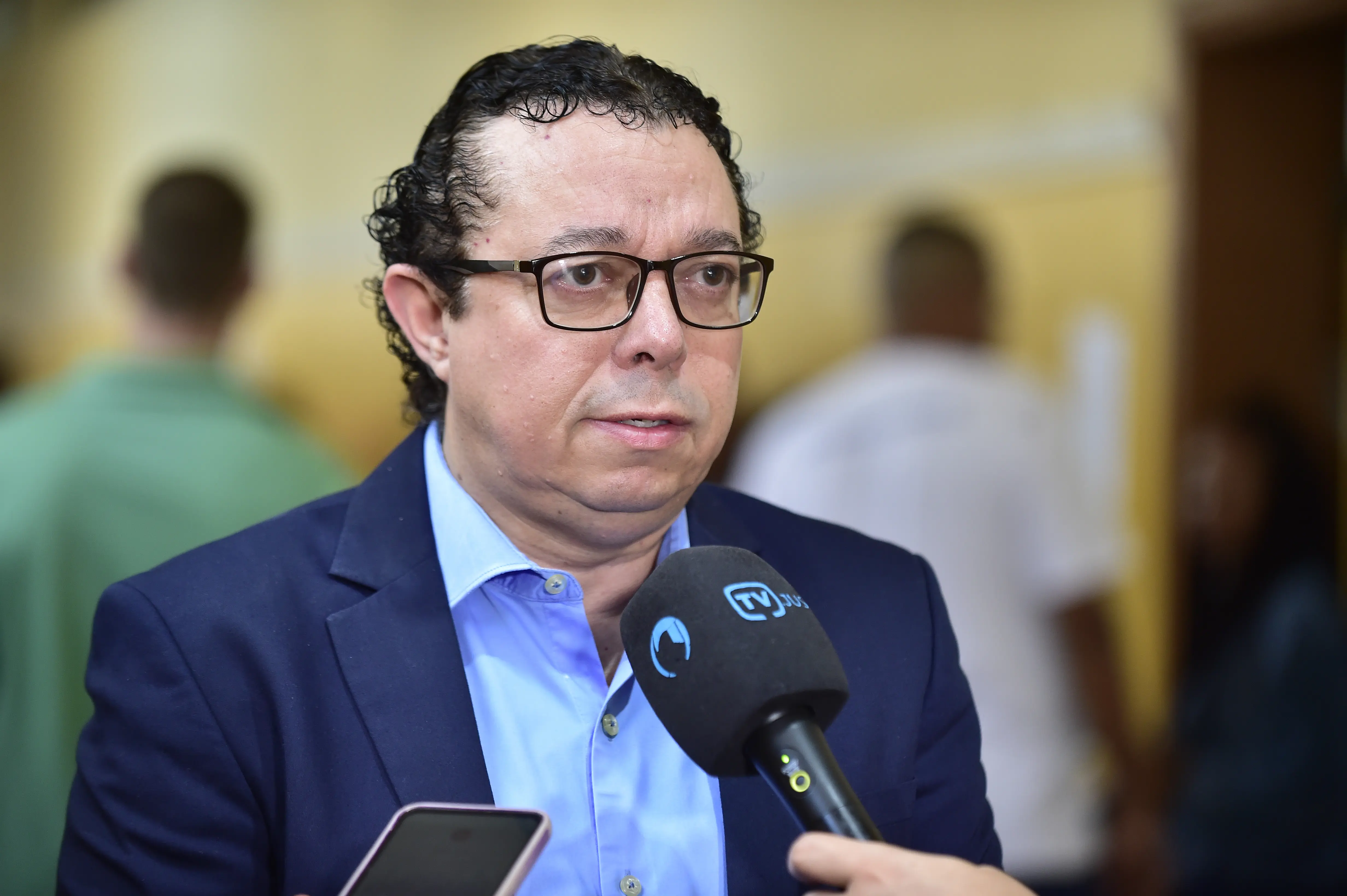 Foto de busto do diretor-geral do TJMT, Flávio de Paiva, concedendo entrevista à TV.Jus. ele é um homem de pele clara, cabelos curtos, pretos e cacheados, olhos castanhos, usando camisa azul clara, terno azul marinho e óculos de grau com armação preta.  