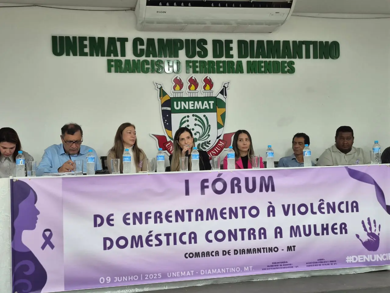 A imagem mostra a mesa de honra do evento, onde estão acomodadas seis pessoas (quatro mulheres e dois homens). À frente, um banner roxo anuncia o "I Fórum de Enfrentamento à Violência Doméstica Contra a Mulher".