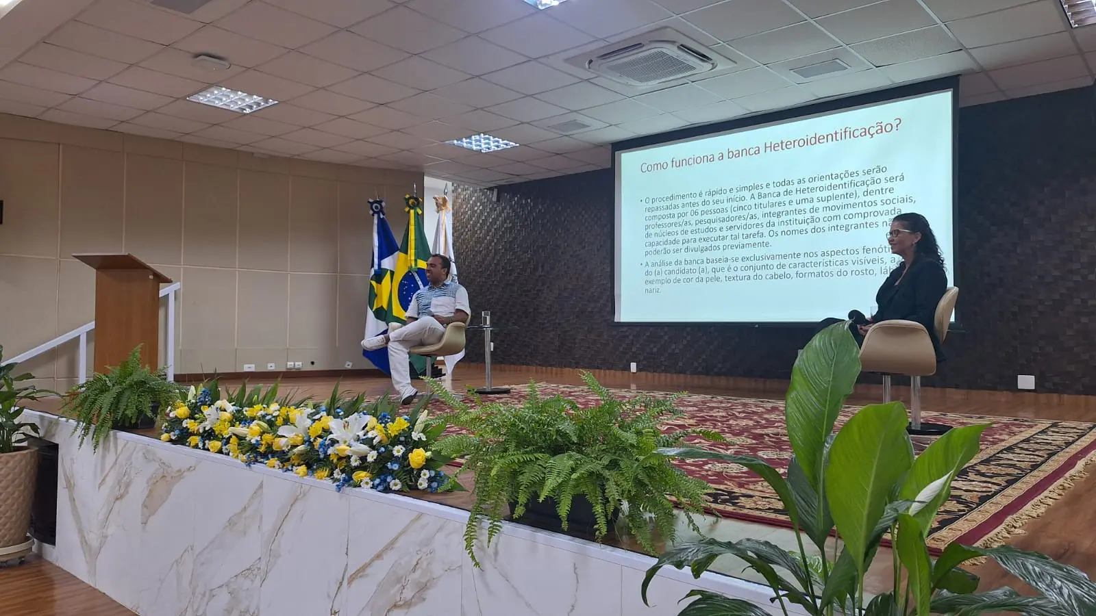 Foto em plano aberto que mostra a palestra sobre bancas de heteroidentificação. No palco estão sentados Manoel Silva, presidente do Conselho Estadual de Promoção da Igualdade Racial, e a presidente da Comissão de Defesa da Igualdade Racial da OAB-MT, Roberta de Arruda. 