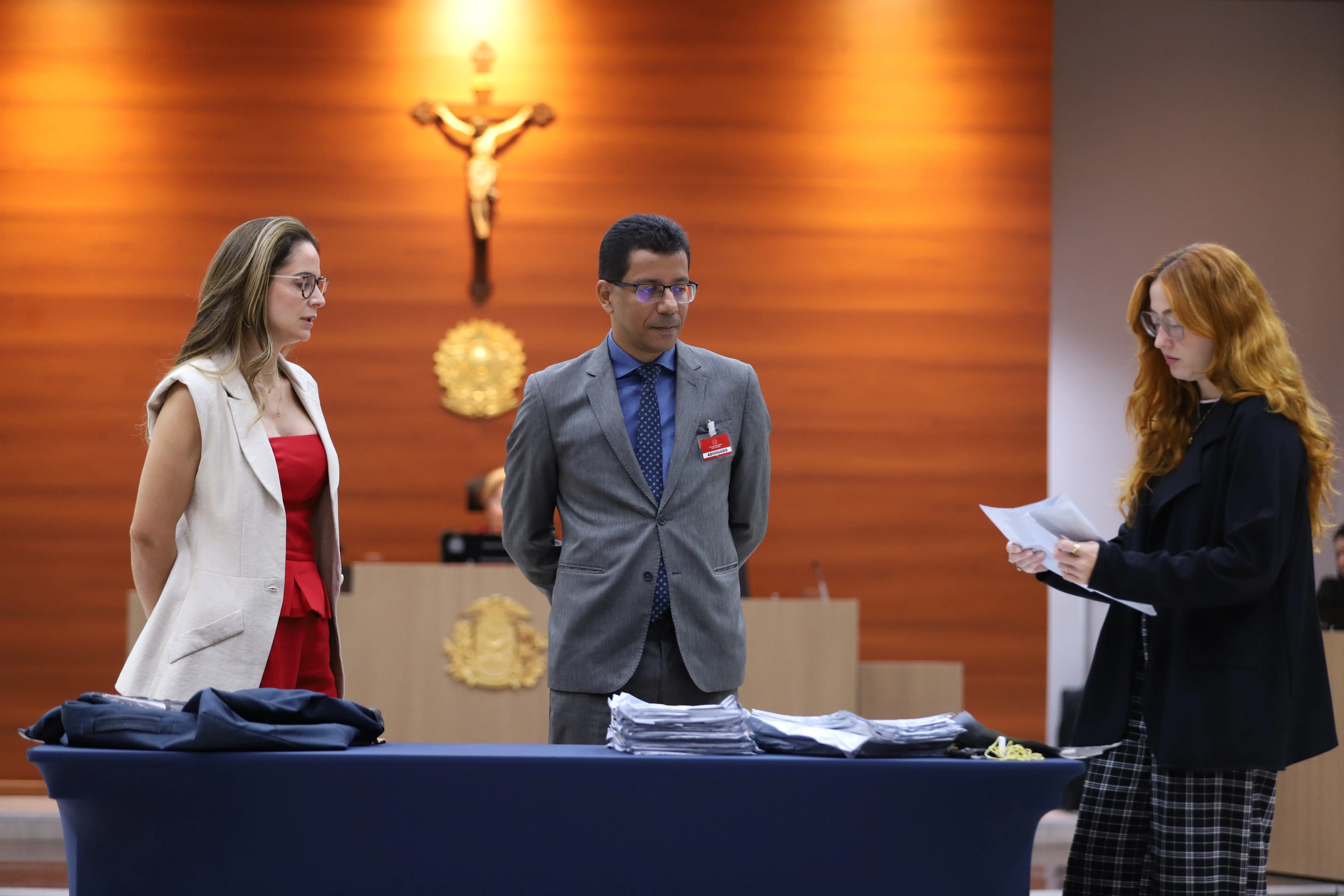 Uma mulher de vermelho e creme, um homem de terno cinza e uma mulher ruiva de preto e xadrez estão em pé ao redor de uma mesa azul, que contém pilhas de papéis e envelopes.